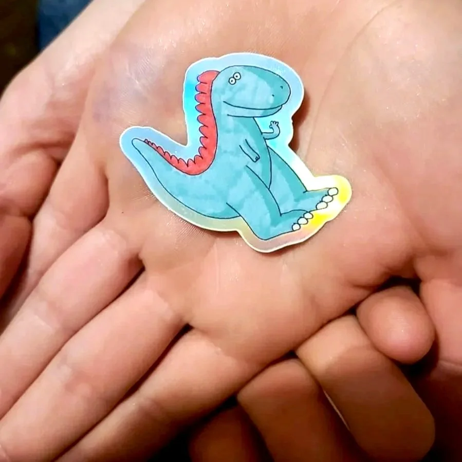 A hand holding a sticker of a blue cartoon dinosaur with red spikes on its back, sitting on a person's finger.
