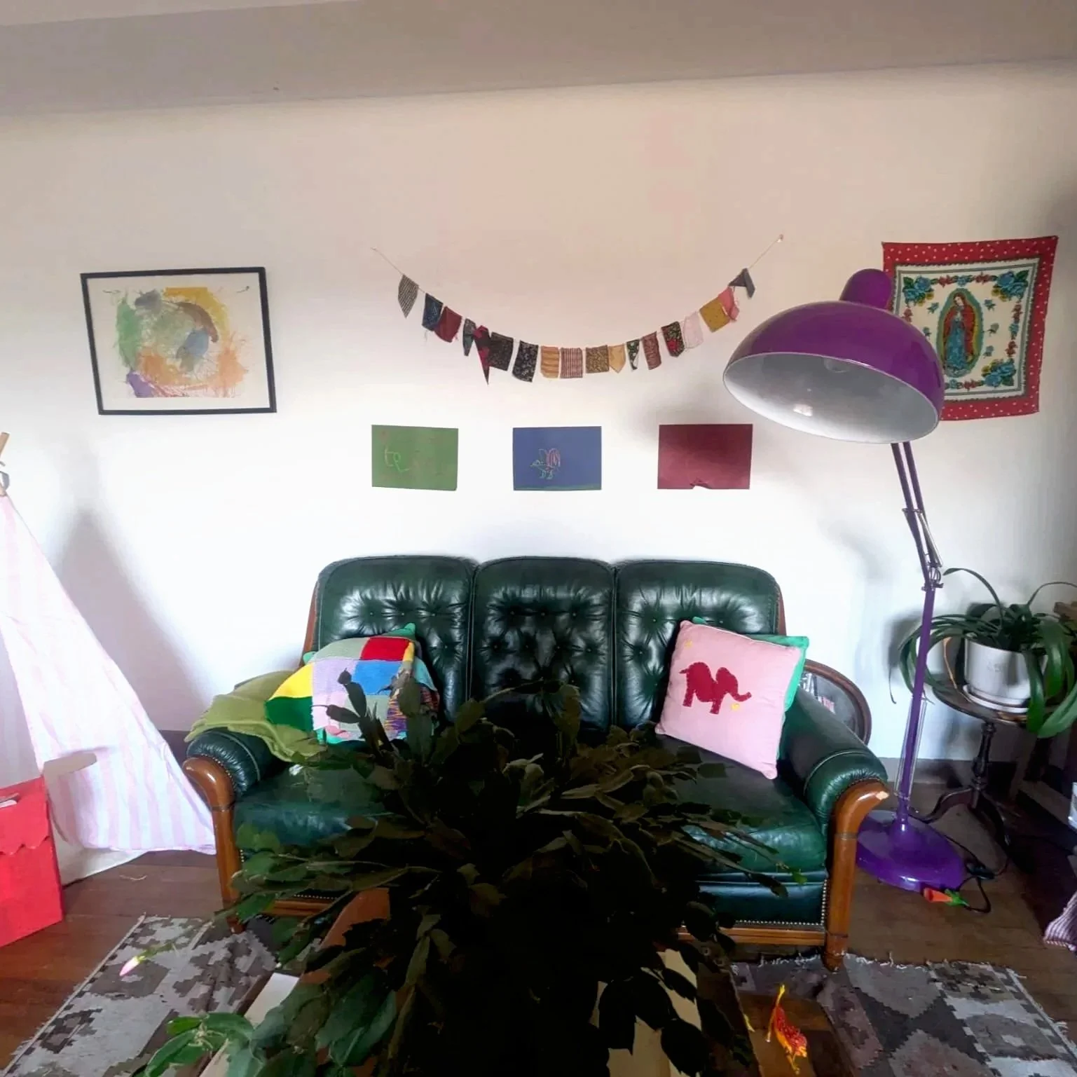 Living room with green leather couch, colorful pillows, potted plant, purple floor lamp, wall art, and decorative garland hanging on the wall.