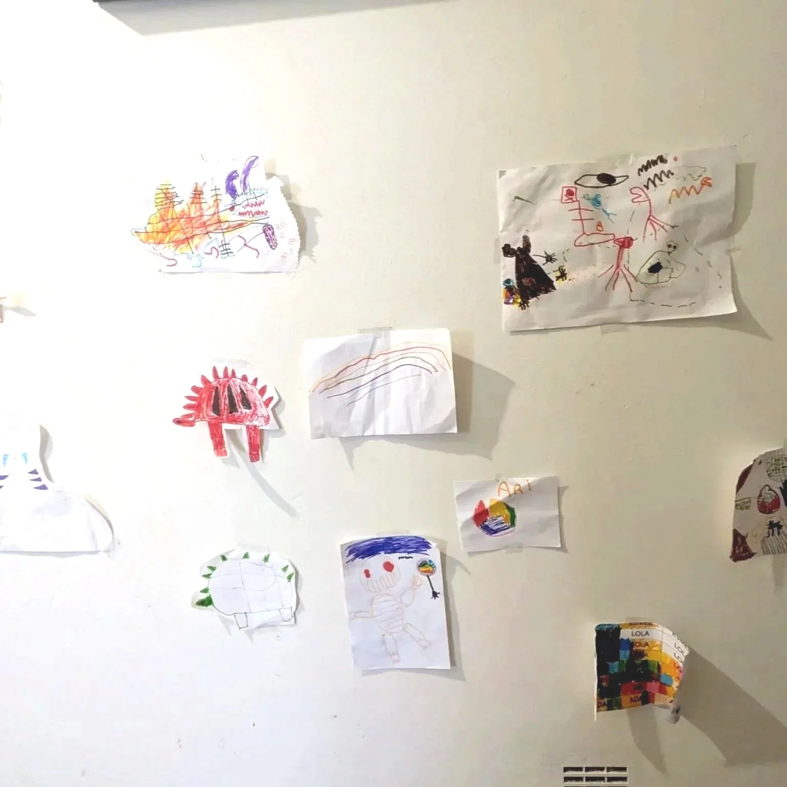 Collection of children's drawings taped to a white wall, including various colorful illustrations of animals, people, and abstract shapes.