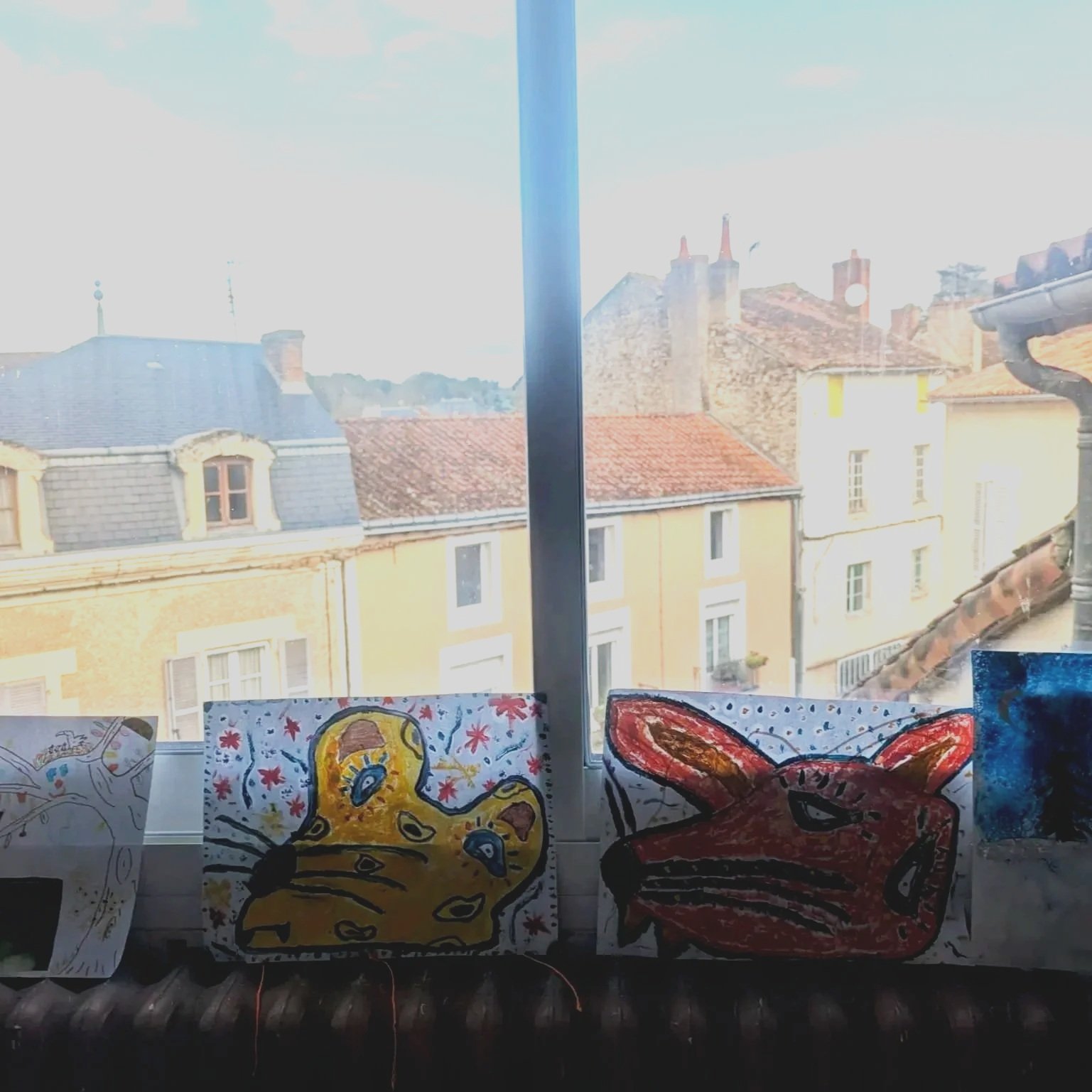 Colorful drawings of animals, including a yellow cat and a red rabbit, displayed on a windowsill with a cityscape of rooftops and chimneys in the background.
