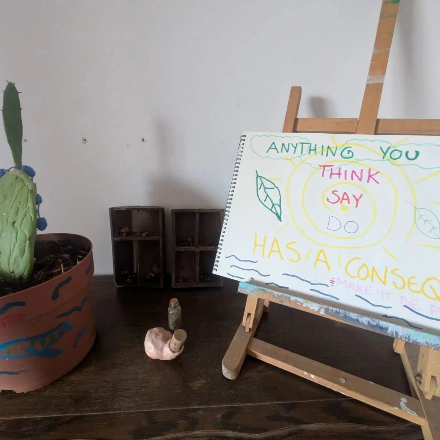 A table with a potted cactus on the left, small empty wooden cubbies behind it, and a sign on an easel on the right with colorful handwritten text and drawings that says: "Anything you think say do has a cause. Make it be positive."