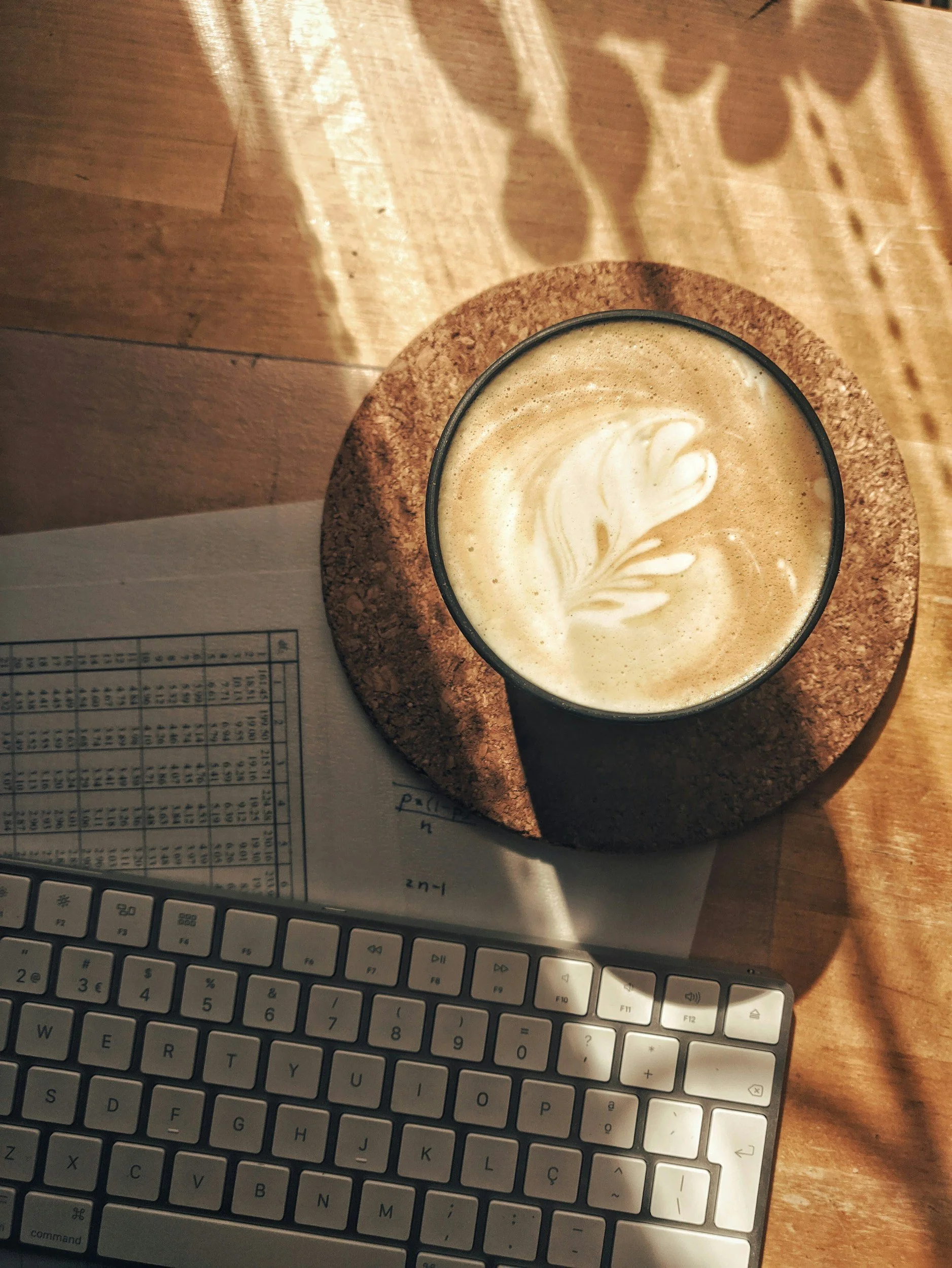 A cup of coffee with latte art on top, placed on a round cork coaster on a wooden desk. Part of a keyboard and a sheet of paper are visible below the coaster.