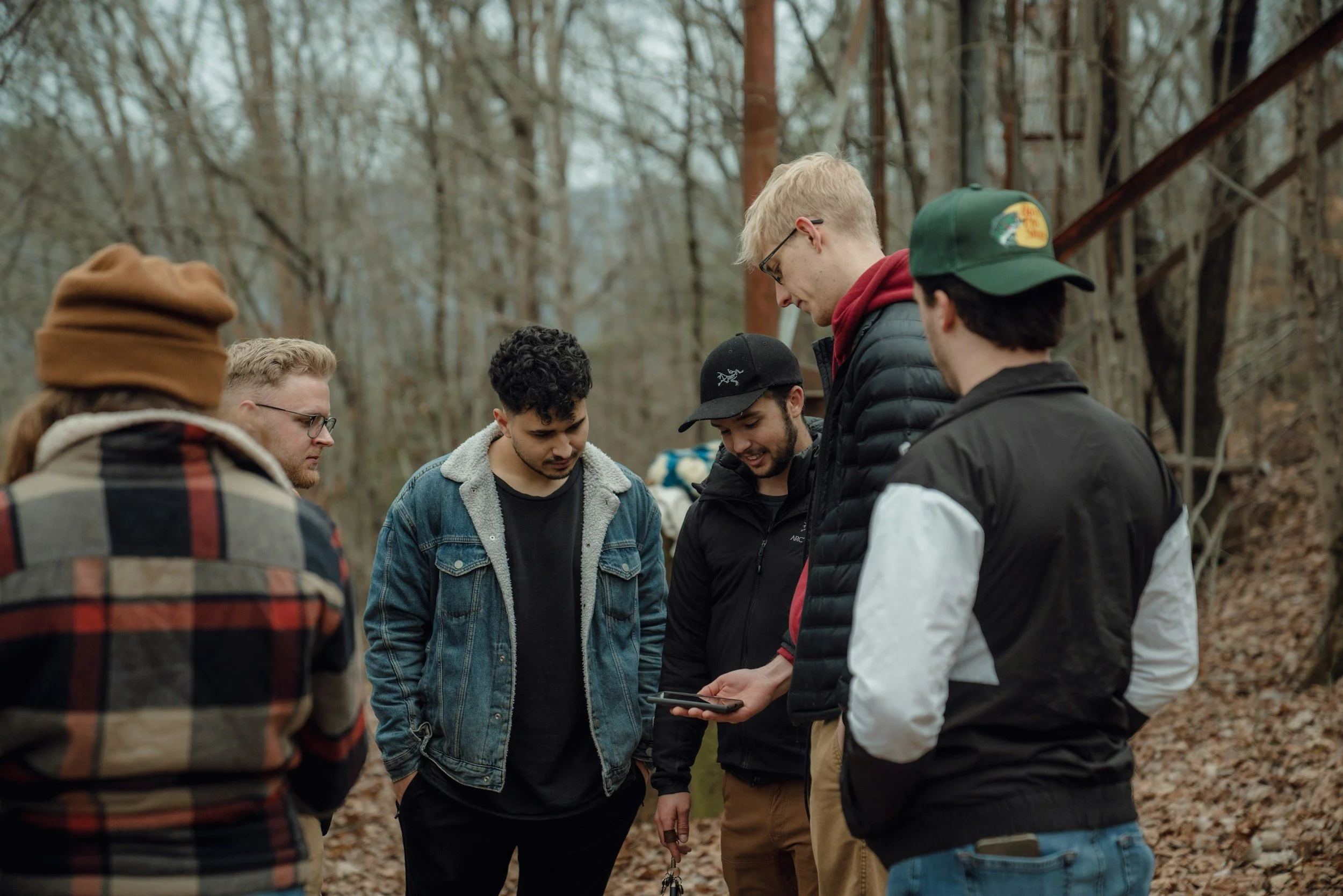 Group of six young men standing outdoors in a wooded area, looking at a smartphone held by one of them.