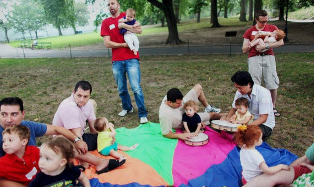 Group of adults and children outdoors on a colorful blanket, some playing drums, others sitting or standing, under trees in a park.