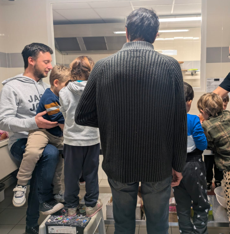 Group of children and adults gathered in a kitchen or laboratory, some standing and some sitting on a small platform, engaged in an activity or demonstration.