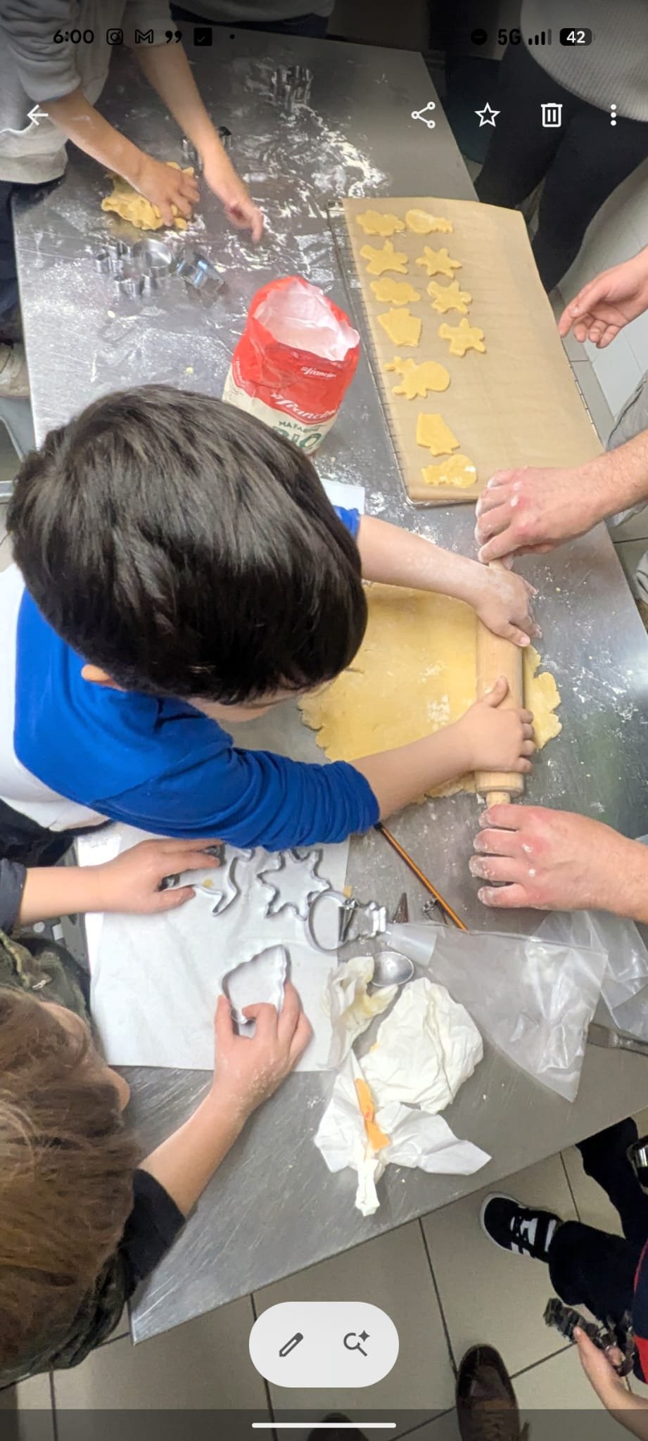 Children and adults rolling out cookie dough on a kitchen table; cookie cutters are nearby and some cookies shaped and cut to be baked.