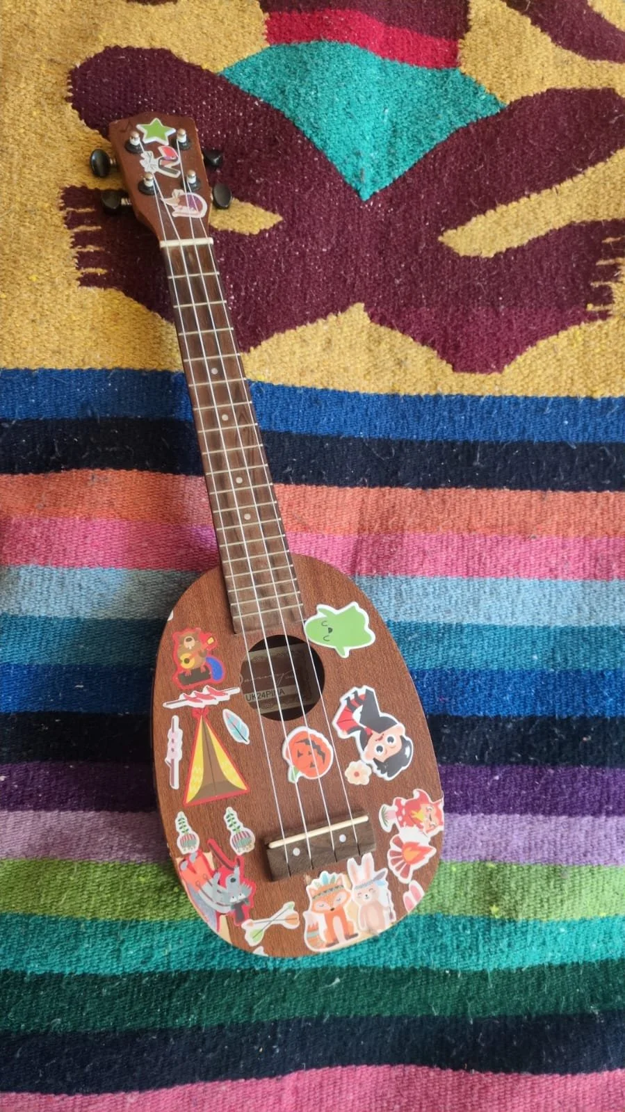 Small wooden ukulele decorated with colorful cartoon stickers, laying on a multicolored striped rug.