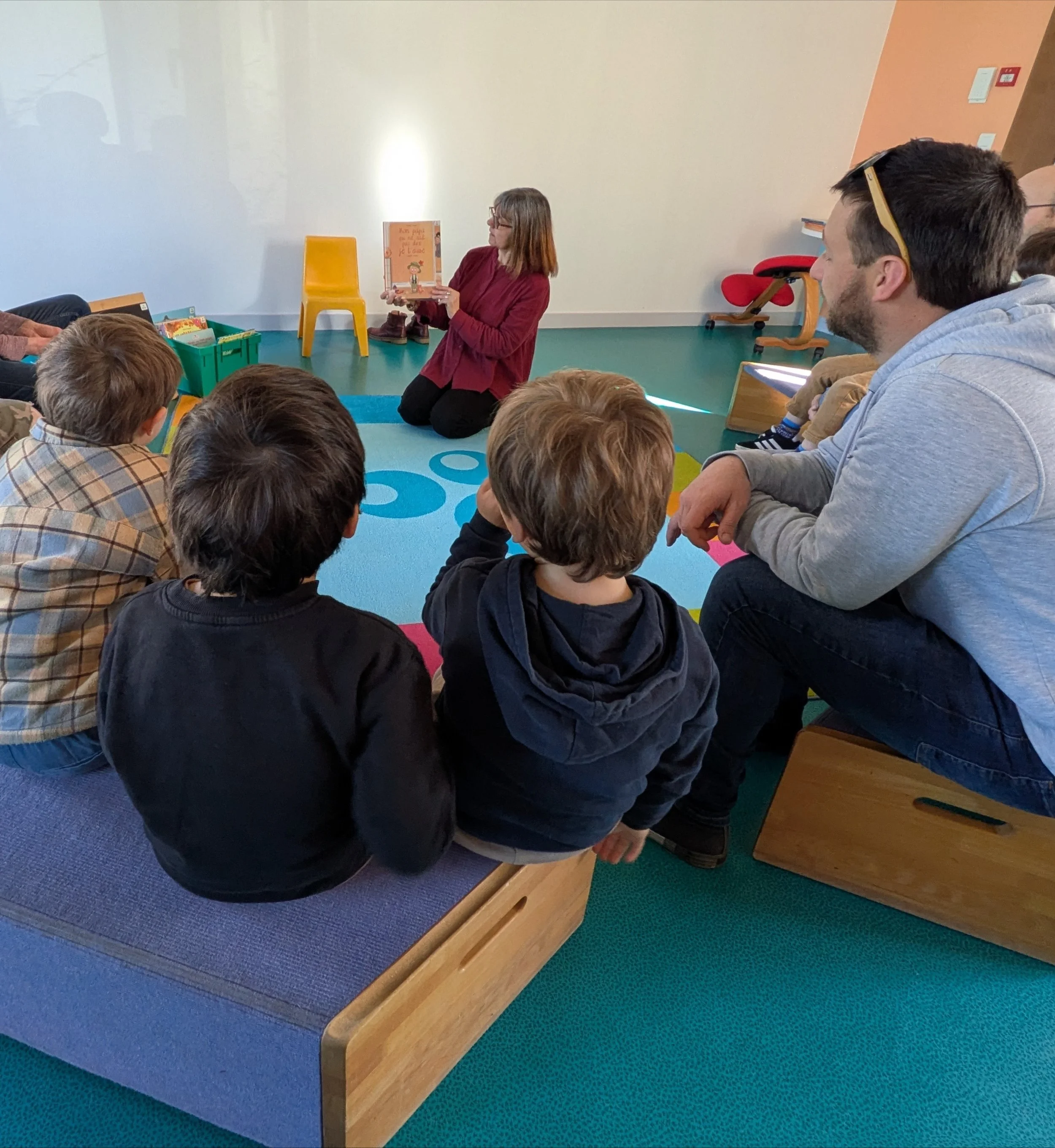 A woman kneeling on the floor reading a book to a group of children seated around her on a colorful playmat in a classroom.