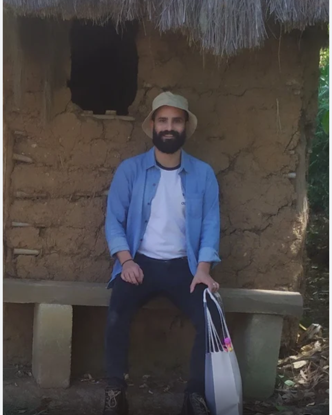 A man with a beard wearing a beige bucket hat, a blue shirt, and black pants sitting on a concrete bench in front of a small mud house with a thatched roof.