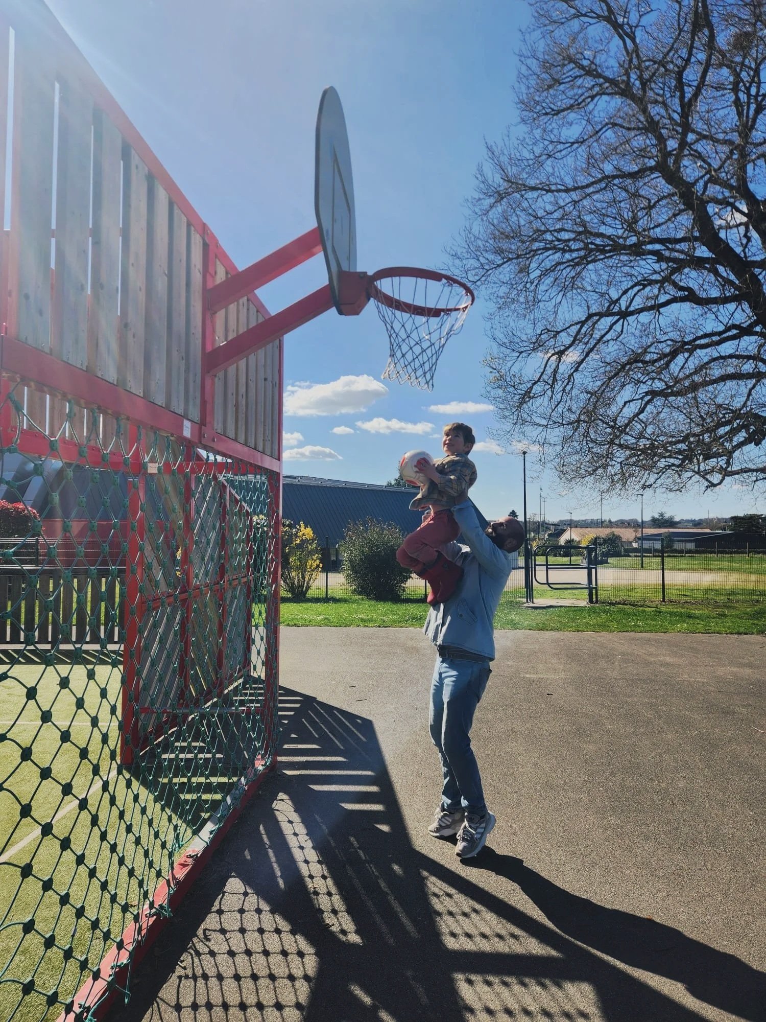 A man is lifting a young boy holding a basketball and a volleyball near an outdoor basketball hoop under a clear blue sky with trees and a fence in the background.