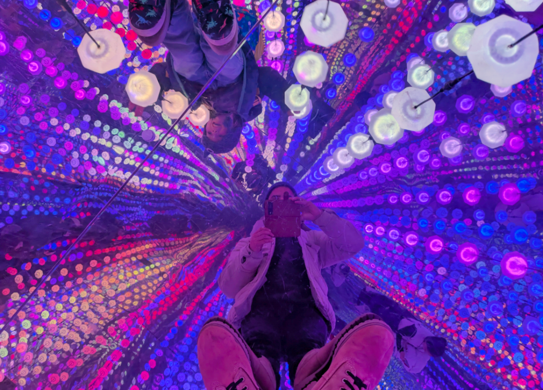 Person taking a mirror selfie inside a colorful, illuminated tunnel with vibrant purple, pink, and blue lights reflecting off mirrored walls.