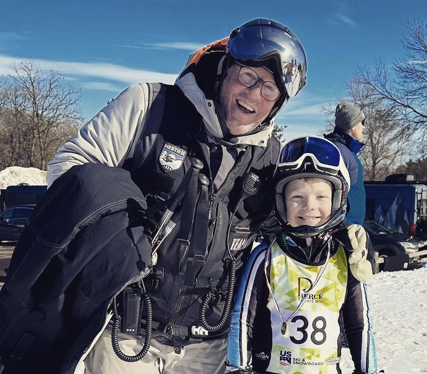 A man and a boy smiling at a snowy outdoor location, both wearing ski helmets and gear, with several trees and vehicles in the background.