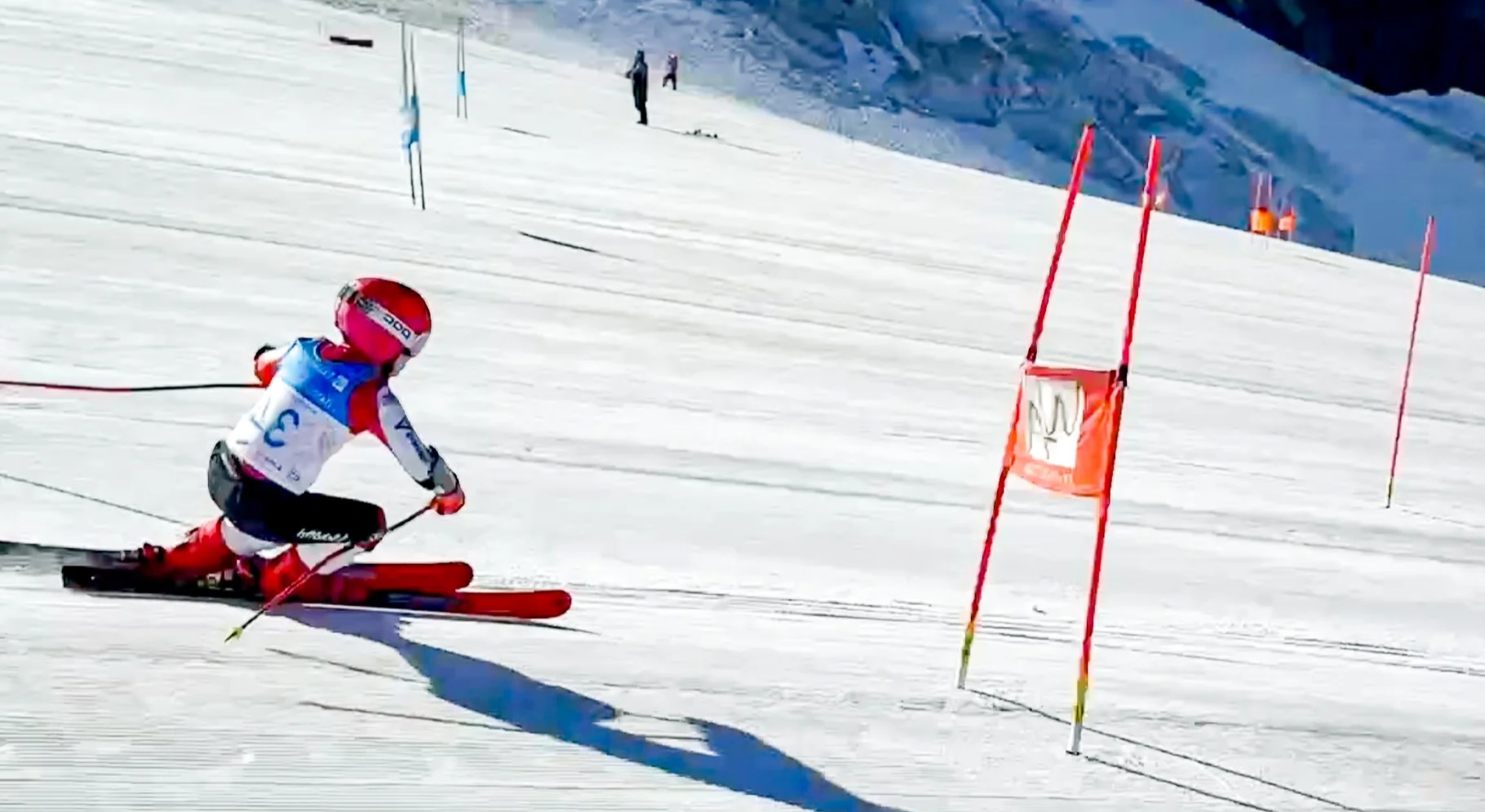 A young skier wearing a pink helmet and a blue and white racing suit navigates through a slalom gate on a snow-covered mountain slope during a ski race.