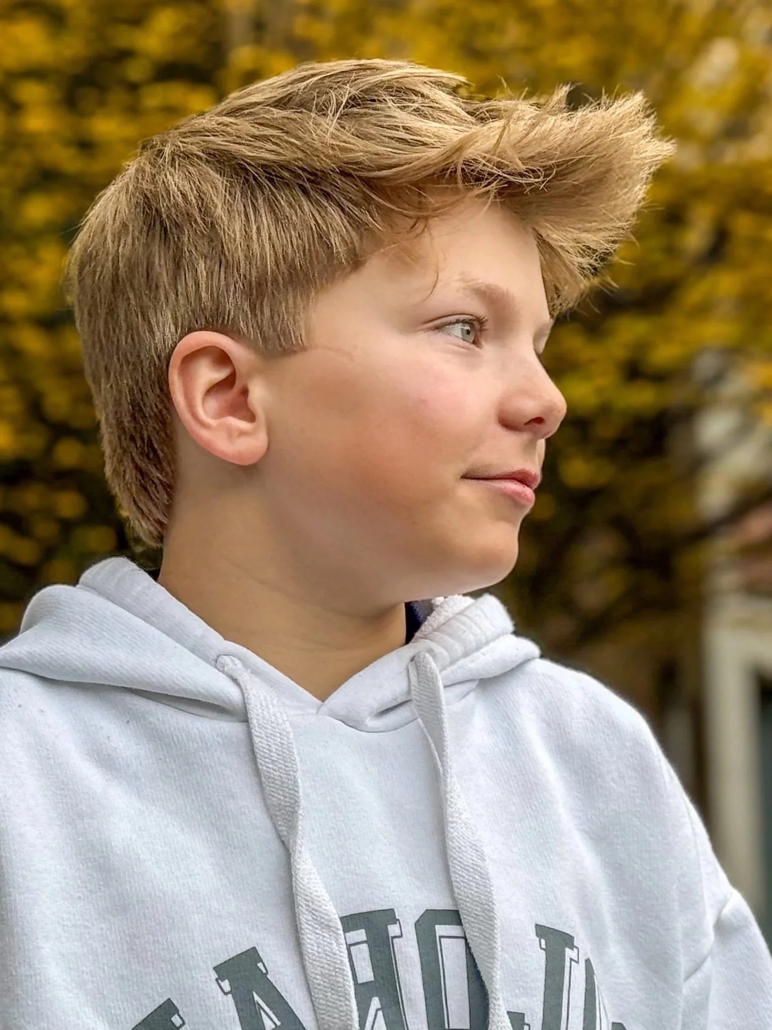 A young person with short, tousled, blonde hair wearing a light gray hoodie, looking to the right outdoors with blurred autumn trees in the background.