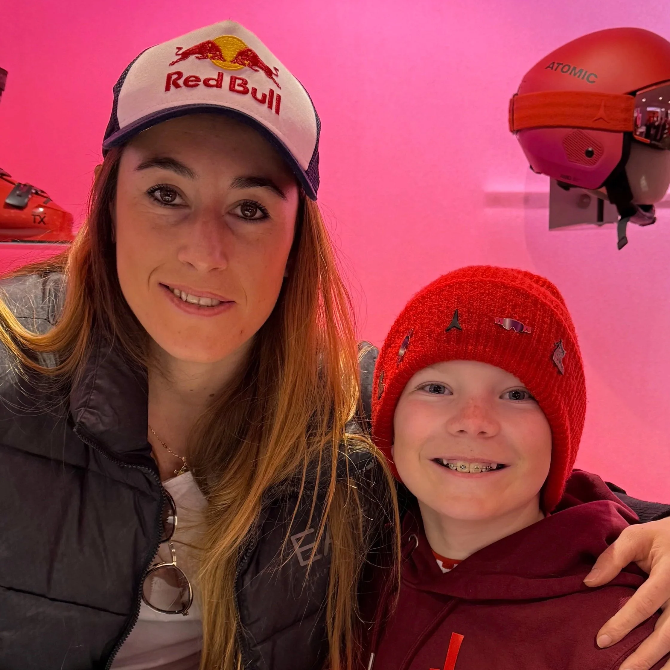 A woman and a young boy taking a selfie together against a pink background with helmets on display. The woman is wearing a Red Bull cap and a black jacket, while the boy is wearing a red knit hat and a maroon hoodie.