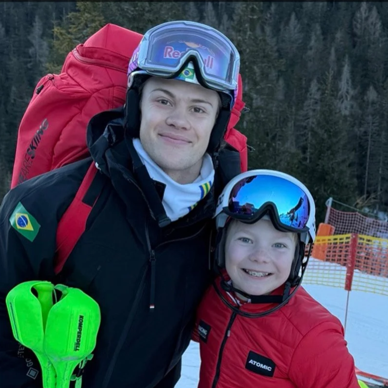 A man and a boy smiling on a snowy ski slope, both wearing helmets and ski gear, with ski goggles on their helmets. The man has a large red backpack and the boy is in a red jacket. Snow fencing and wooded mountains are in the background.