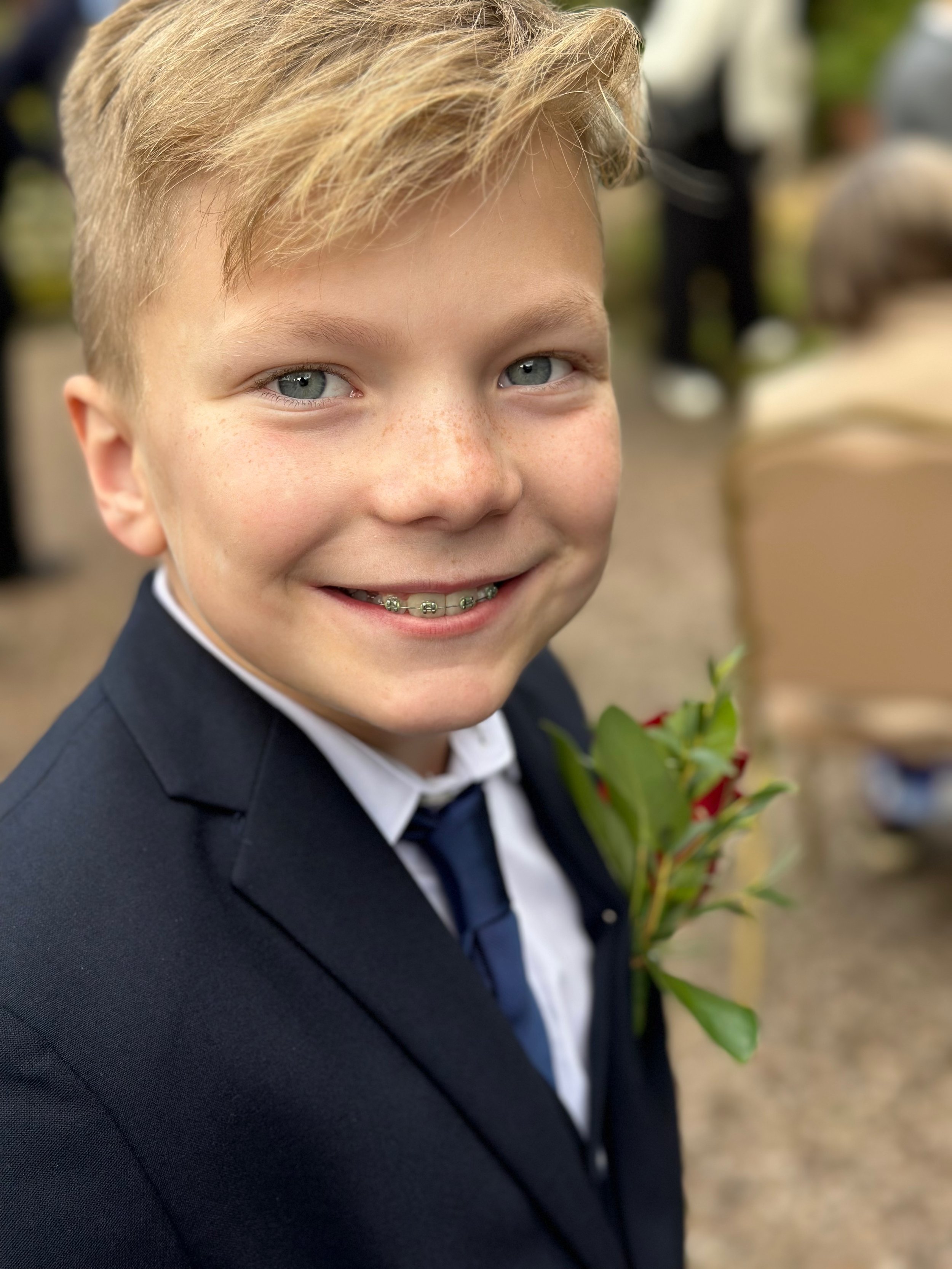 A young boy with blonde hair, blue eyes, and braces smiling outdoors. He is dressed in a dark suit with a white shirt, a tie, and has a flower pinned to his suit.