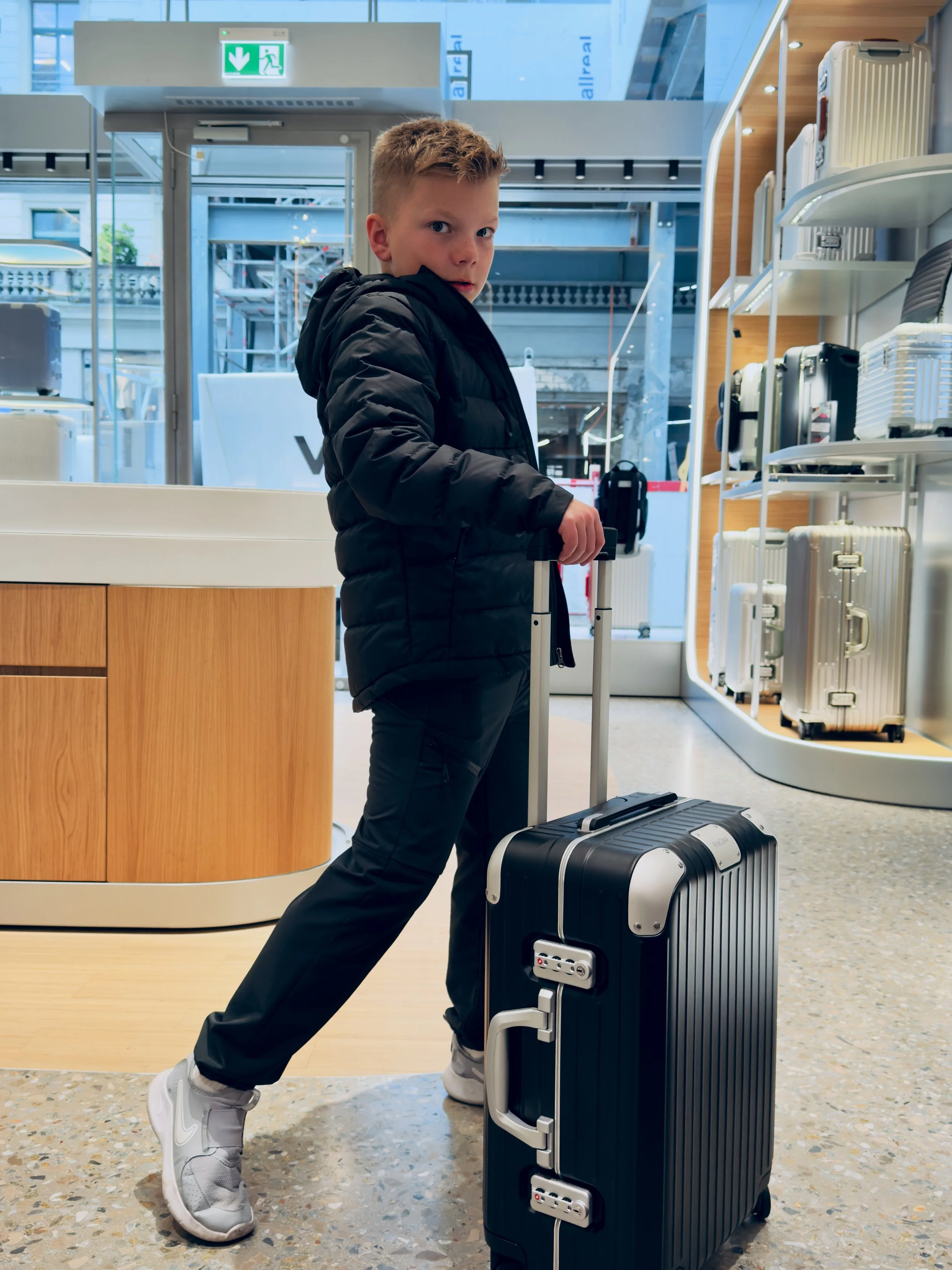 Young boy in black jacket and pants pulls a black rolling suitcase at an airport, standing inside near luggage shelves and glass doors.
