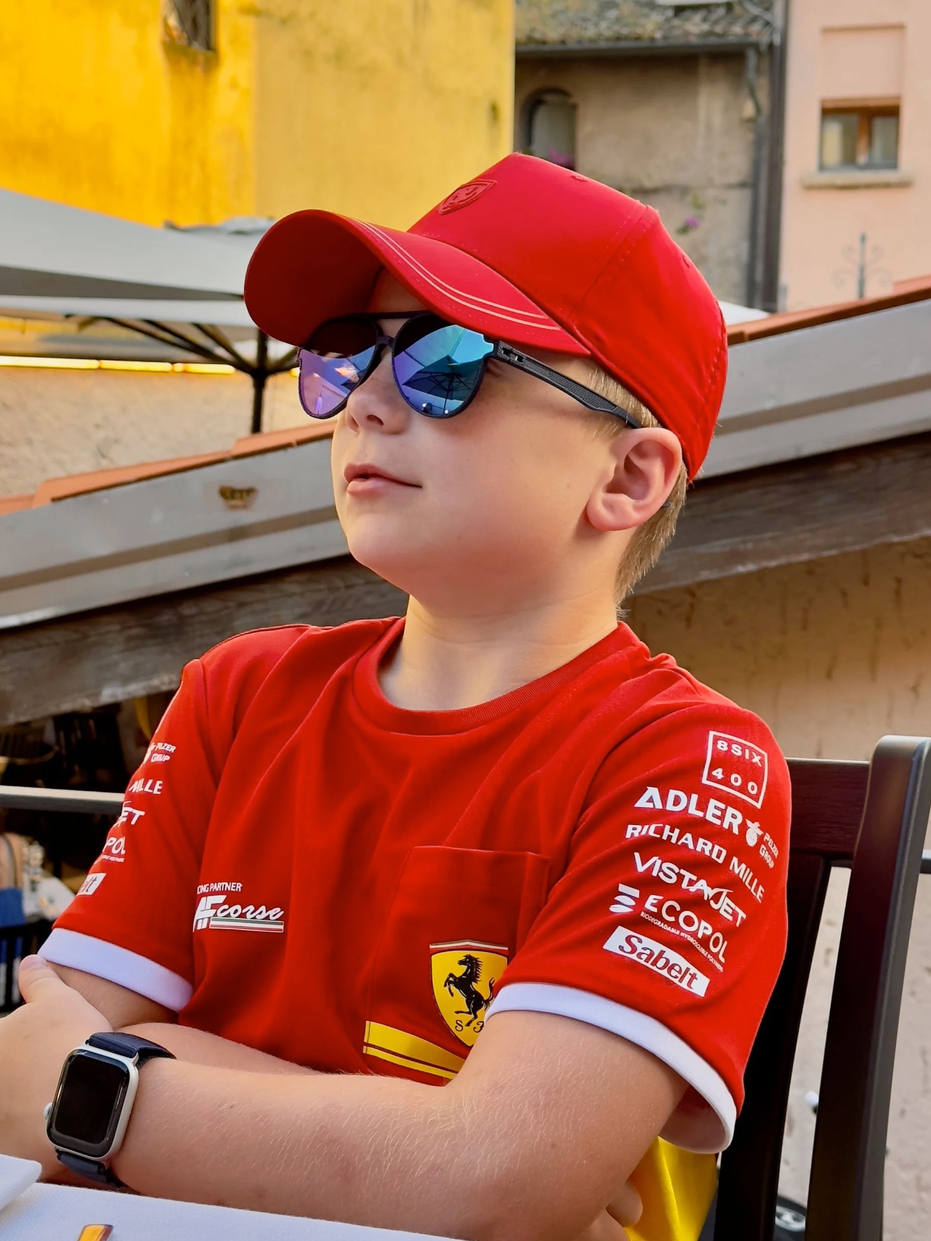 A young boy wearing sunglasses, a red cap, and a red Ferrari racing shirt with sponsor logos, sitting outdoors with arms crossed.