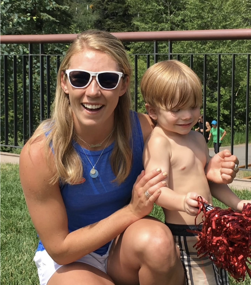 Mikaela Shiffrin,  smiling and sitting on the grass outdoors. A young Daniel Fatzinger is sitting next to her, also smiling. They are in Beaver Creek, Colorado at a holiday event.