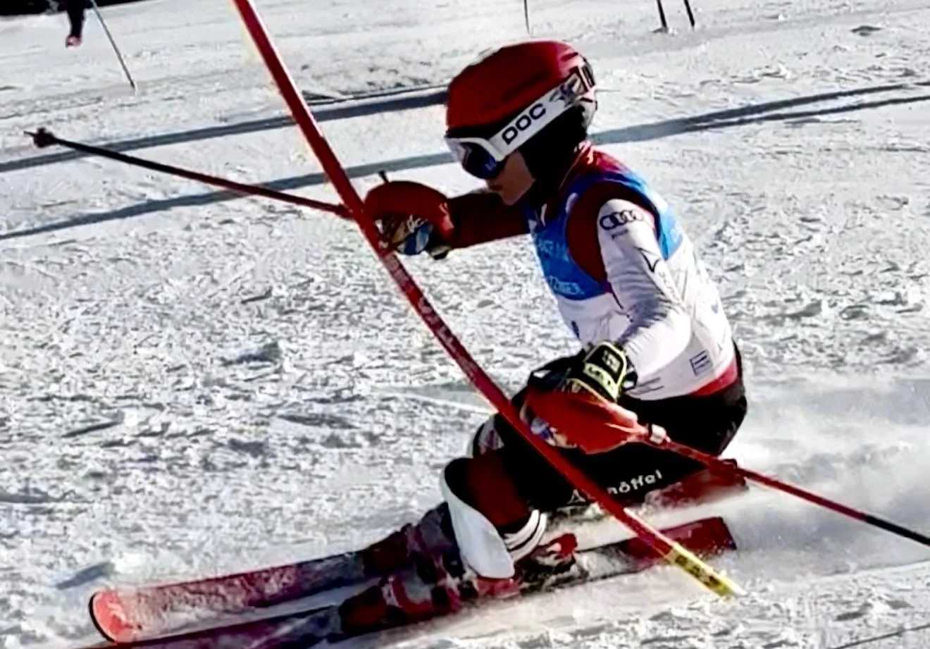 A skier wearing a red helmet, blue and white racing suit, and black gloves kneels on snow beside their red skis and poles, suggesting a skiing competition or training session on a snowy slope.