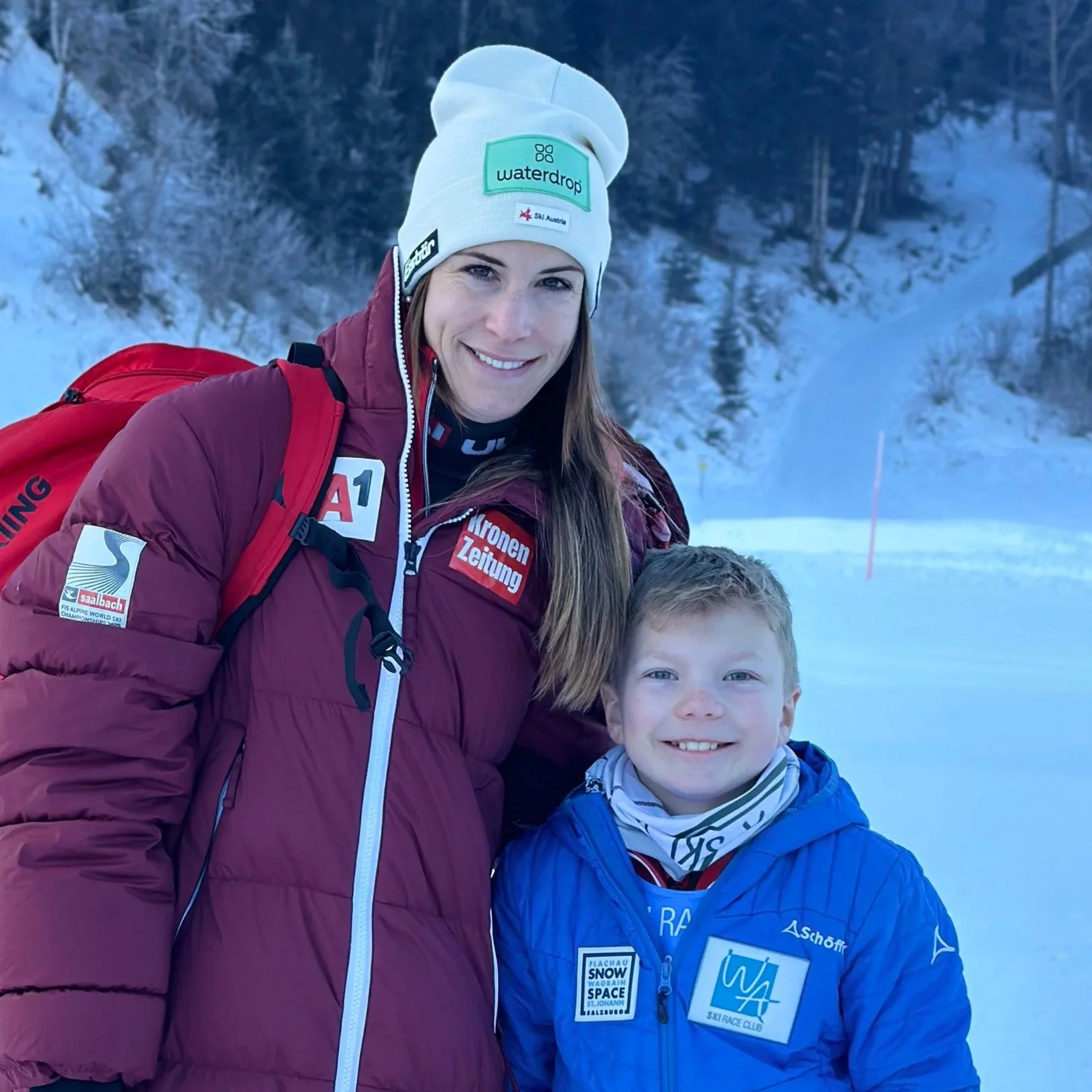 A woman in a maroon jacket and white beanie and a young boy in a blue jacket smiling outdoors in a snowy winter landscape.