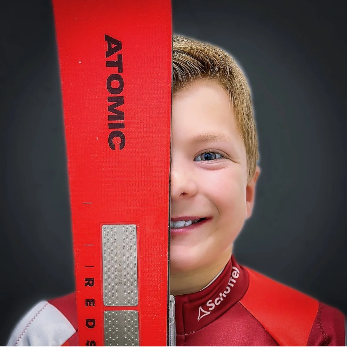 Young male cyclist smiling, holding a red bicycle frame with the brand 'Atomick' visible, wearing a red and white cycling jersey with a logo on the collar, against a dark background.