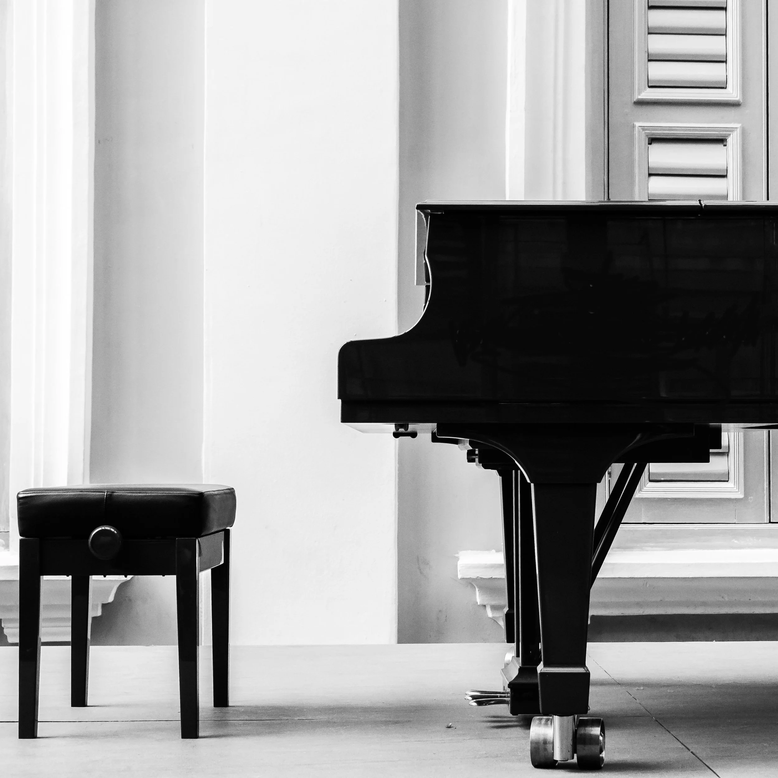 Schwarz-weiße Aufnahme eines Klaviers mit Hocker vor einem Fenster mit Jalousien in einem Raum.