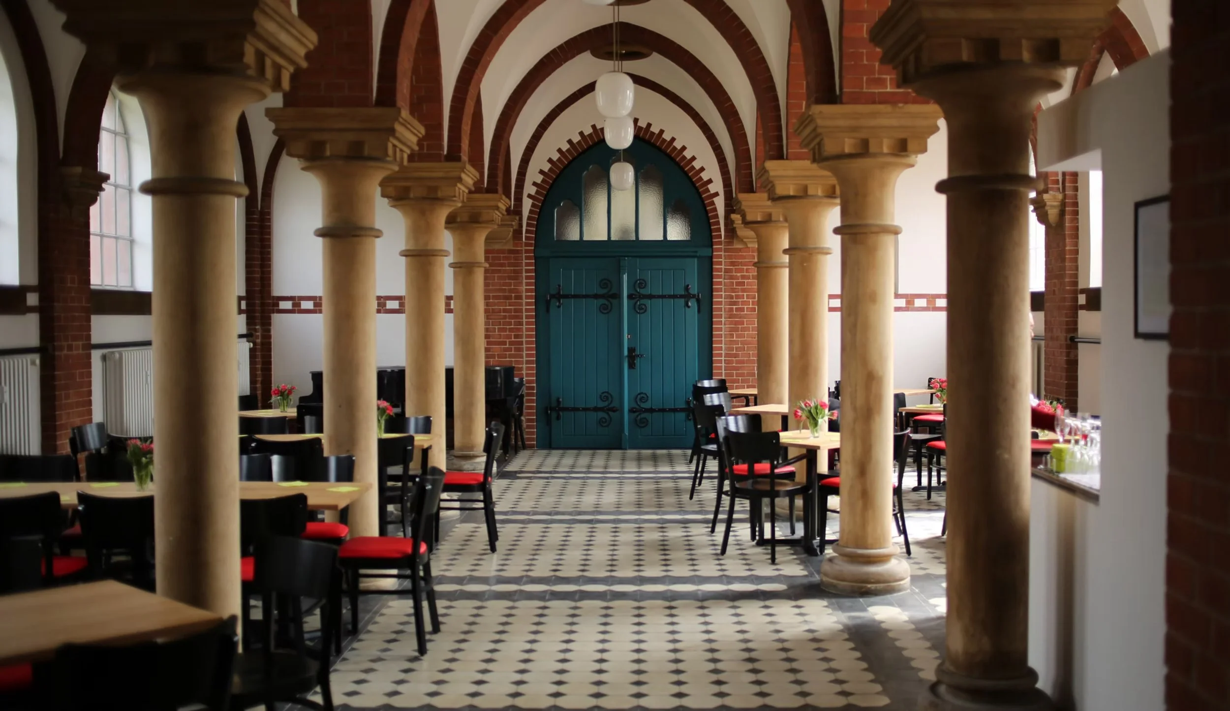 Innenraum eines Restaurants mit Säulen, schwarzen Stühlen mit roten Polstern, Tischen mit Blumen, große Tür mit Bogen im Hintergrund.