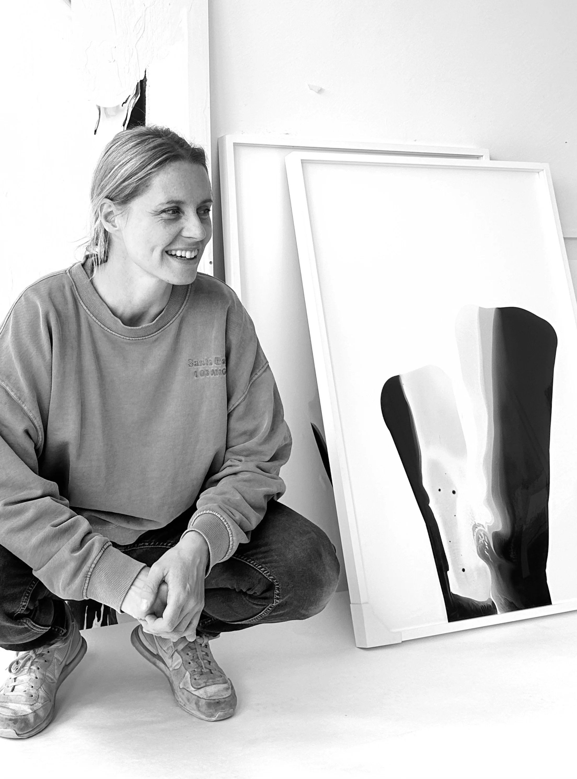 Eine Frau in einem blauen Pullover und Jeans sitzt neben einem weiß gerahmten abstrakten Gemälde mit grünen, weißen, gelben, roten und braunen Farbschichten.