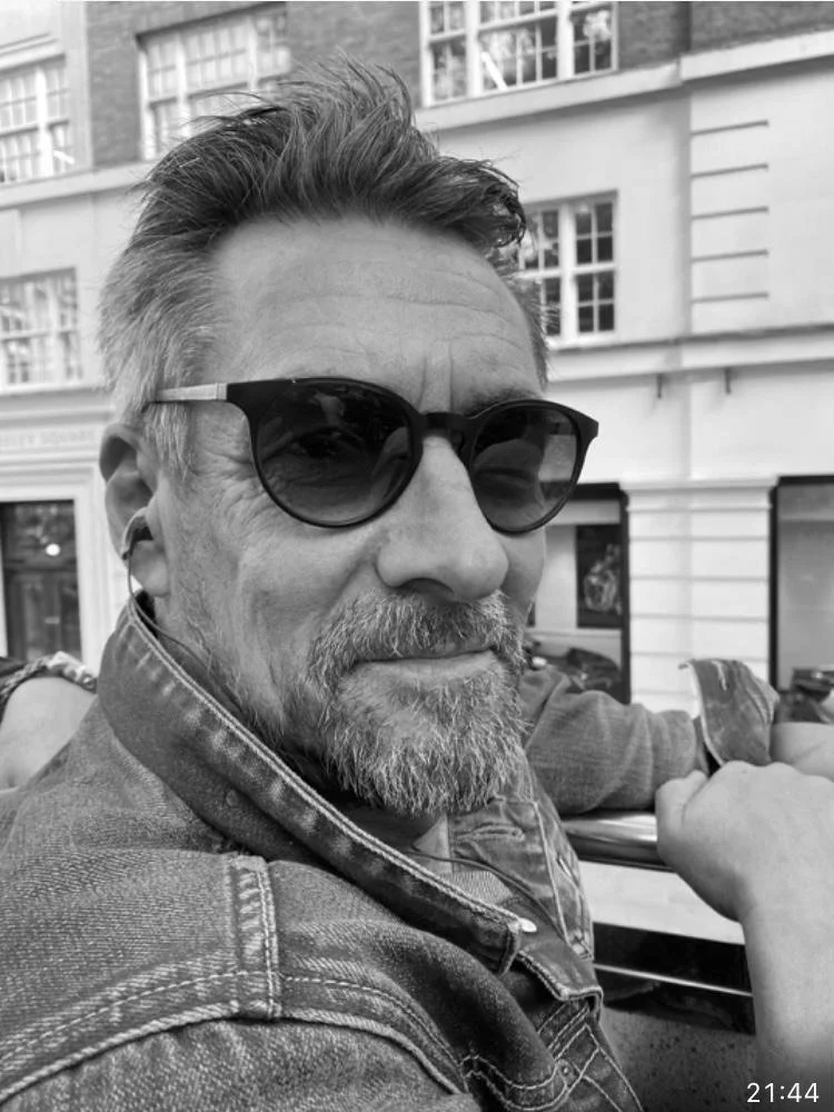 Ein Mann mit grauem Bart, Sonnenbrille und Jeansjacke, lächelnd, draußen in einer Stadt.