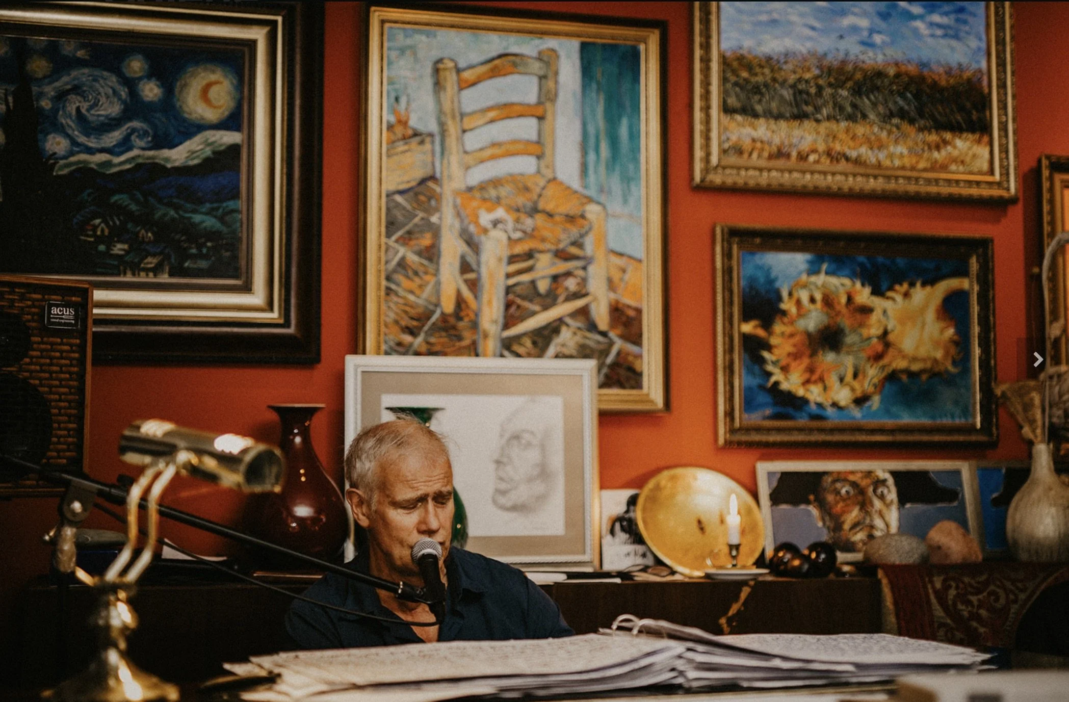 Ein ältere Mann sitzt in einem Raum auf seinem Schreibtisch, im Hintergrund eine Wand voll von Kunstwerken und Gemälden.