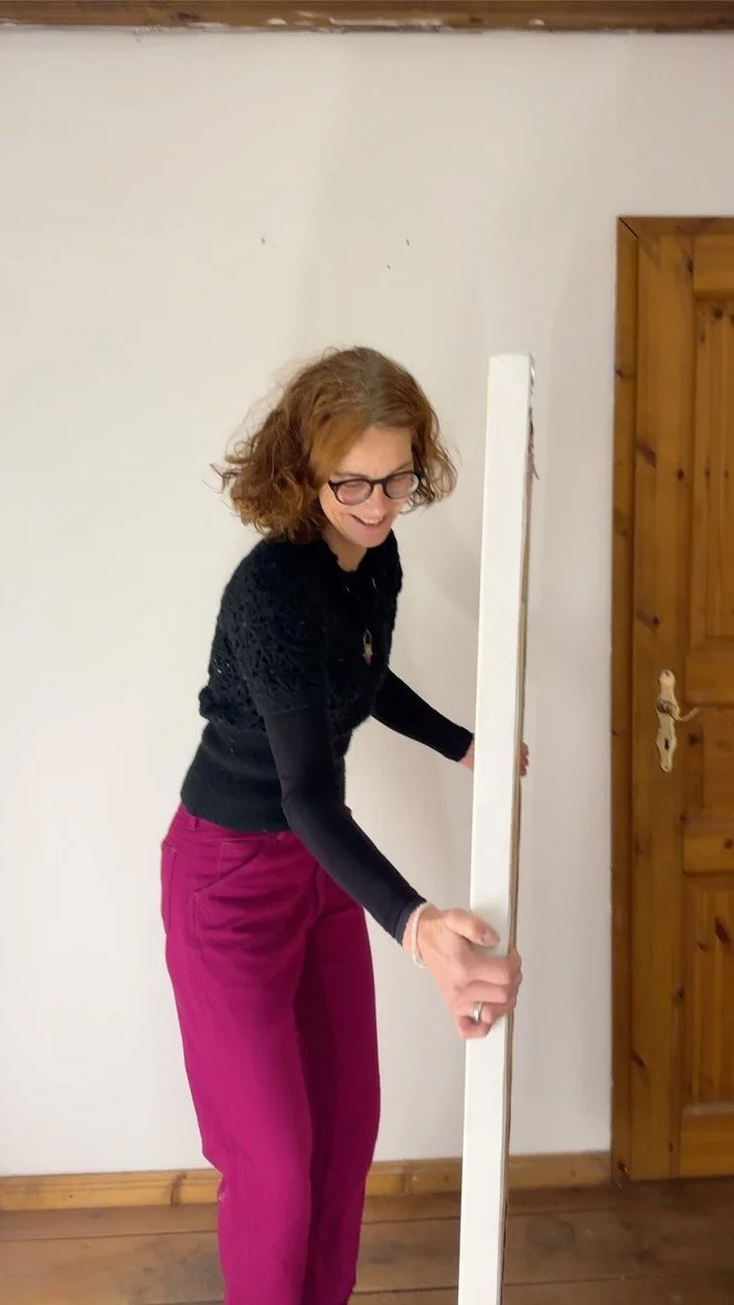 Eine Künstlerin mit Brille namens Simone Westphal und lockigem braunem Haar hält ein Gemälde in der Hand und lächelt, während sie im Raum steht.