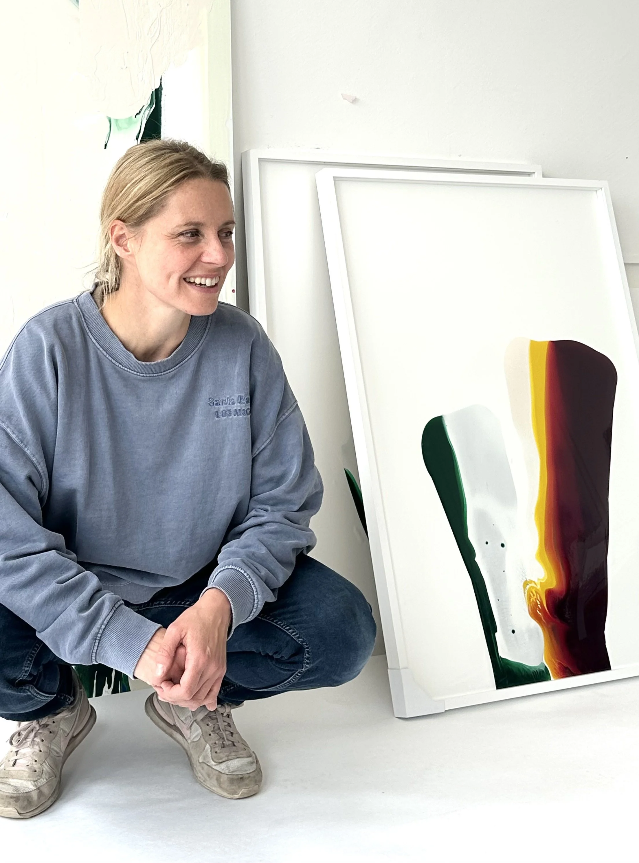 Eine Frau in einem blauen Pullover und Jeans sitzt neben einem weiß gerahmten abstrakten Gemälde mit grünen, weißen, gelben, roten und braunen Farbschichten.