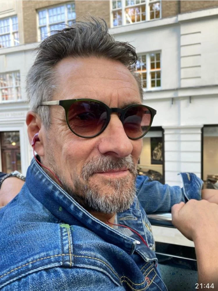 Ein Mann mit grauem Bart, Sonnenbrille und Jeansjacke, lächelnd, draußen in einer Stadt.