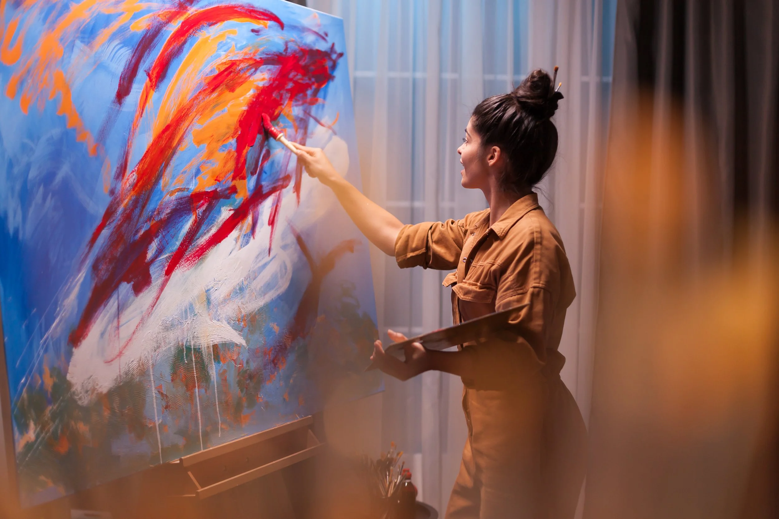 Eine Frau malt abstrakte Kunst auf eine große Leinwand in einem Raum mit Vorhängen.
