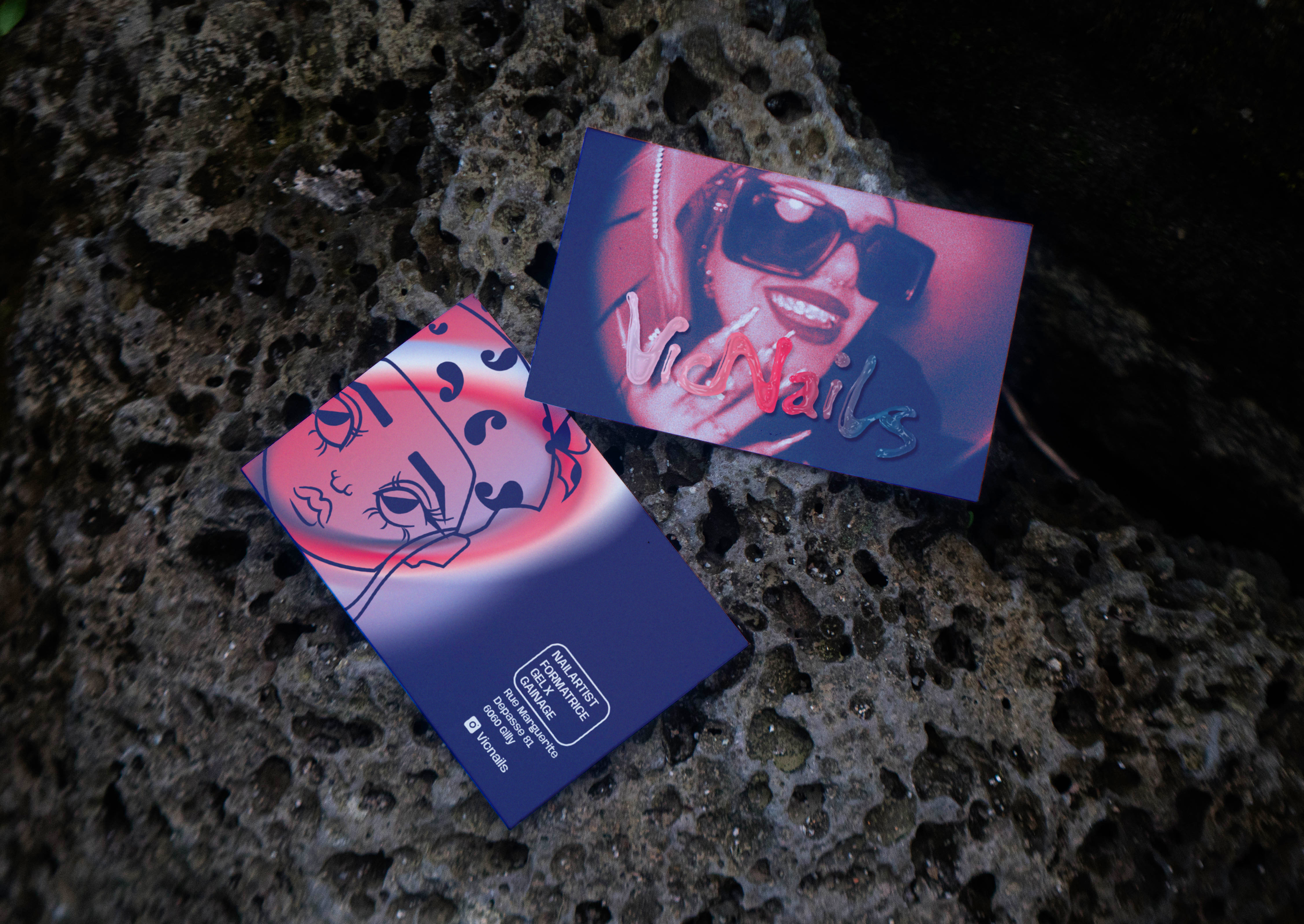 Deux cartes de visite, l'une représentant une femme portant des lunettes de soleil et souriant, avec le mot 'VICNails' inscrit, et l'autre avec une illustration de visages féminins stylisés, posées sur une surface rocheuse.