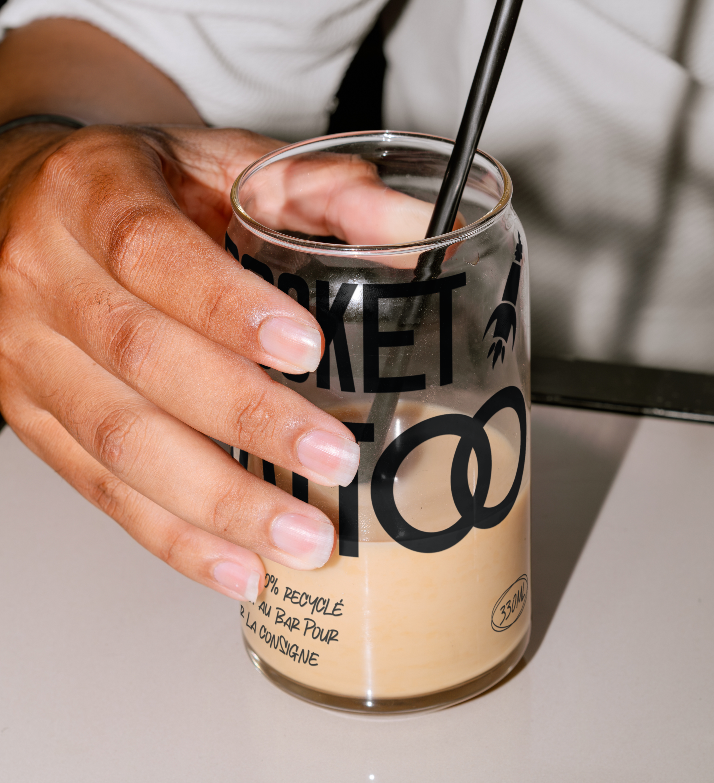 Une main tient un verre de verre avec une boisson beige claire et une paille noire. Le verre a des inscriptions promotionnelles sur la recyclabilité, et une partie de l'arrière-plan montre une surface claire et un vêtement à rayures blanc et noir.