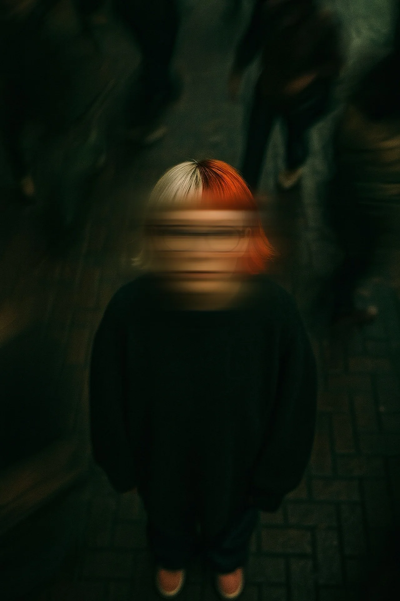 Une personne debout vue de dessus, portant un sweatshirt noir, avec des cheveux courts mi-rouges, mi-blancs, devant un mur sombre. L'image est floue au niveau du visage.