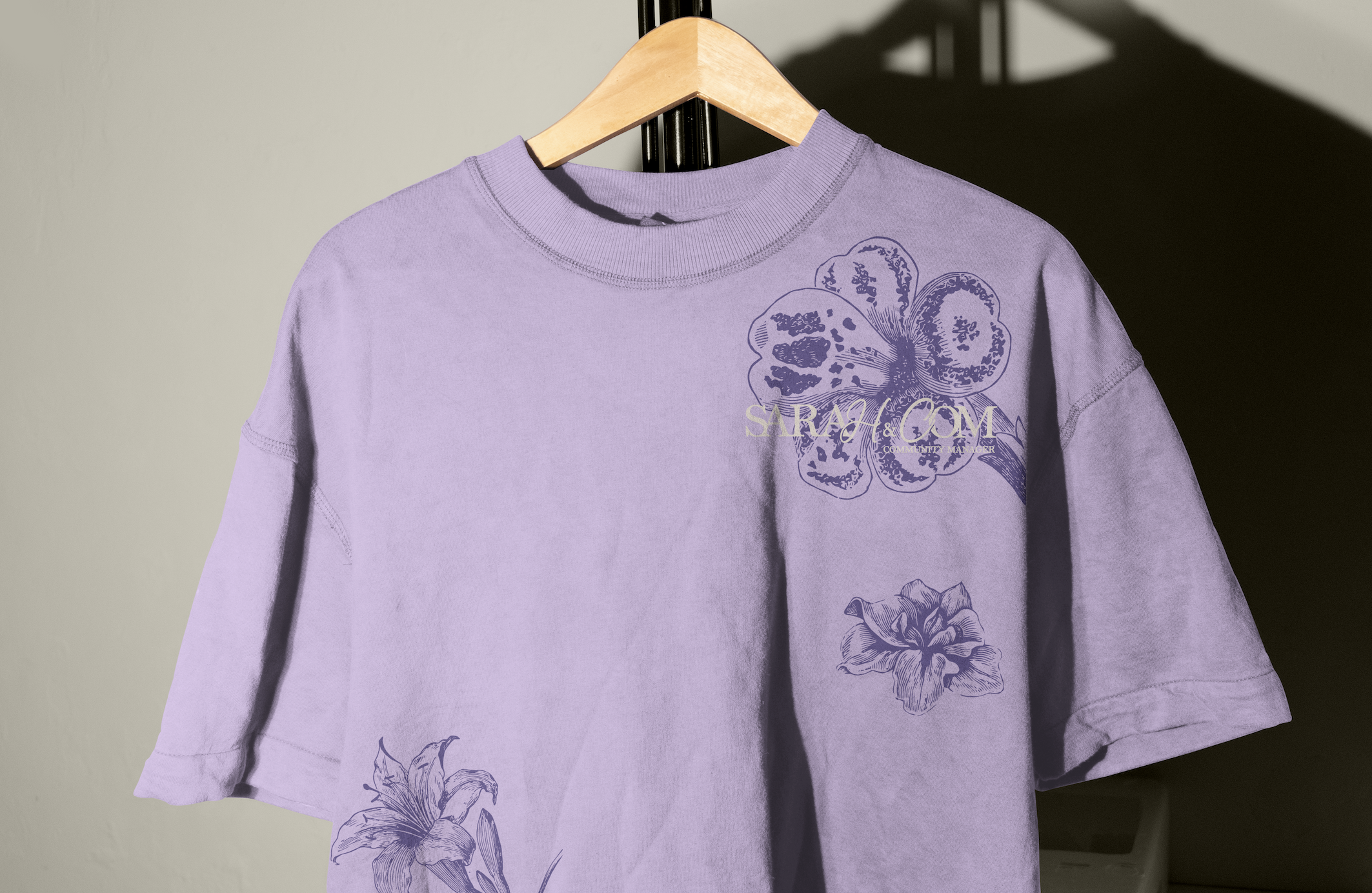 Un t-shirt violet avec des motifs floraux en haut à droite et en bas à gauche, suspendu sur un cintre en bois, avec un fond en lumière et une ombre portée noire.