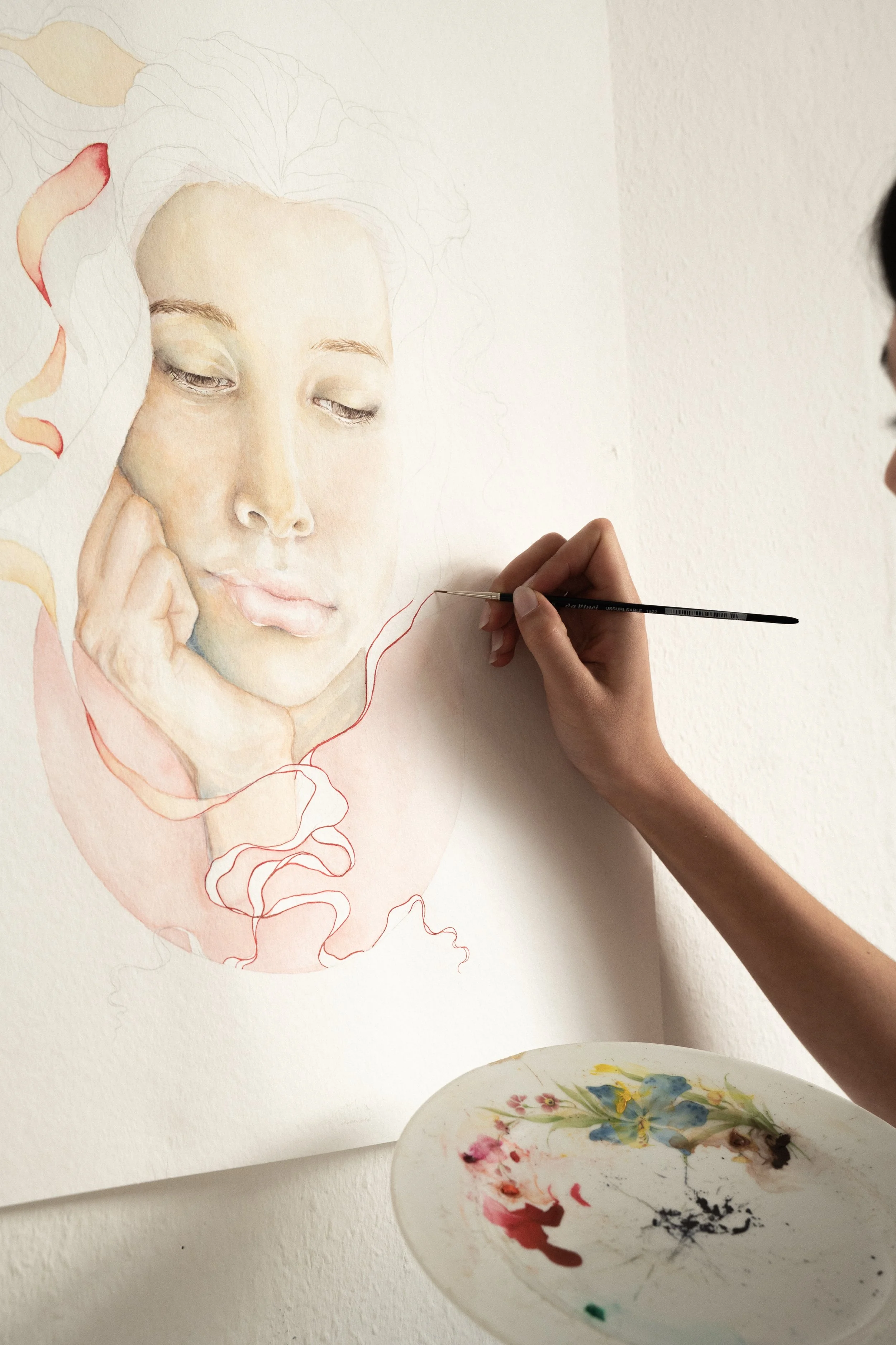 Künstler malt ein realistisches Porträt einer jungen Frau auf Papier, während sie die Details mit einem Pinsel ausbessert, mit einer Farbpalette auf dem Tisch.