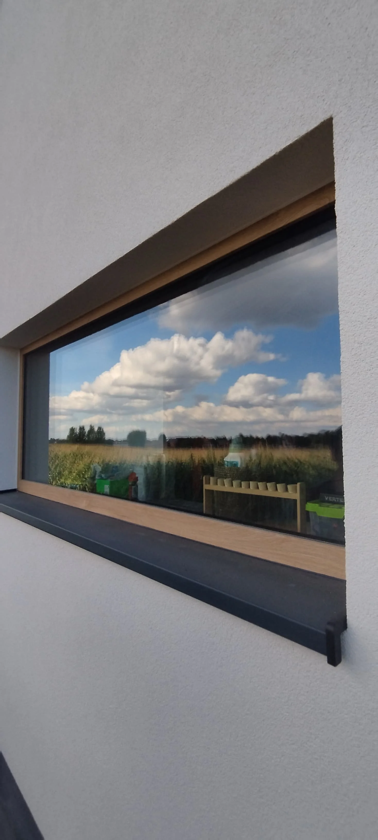 Uitzicht door een raam met een houten raamkozijn op een landschap met een grash田d en een blauwe lucht met witte wolken.