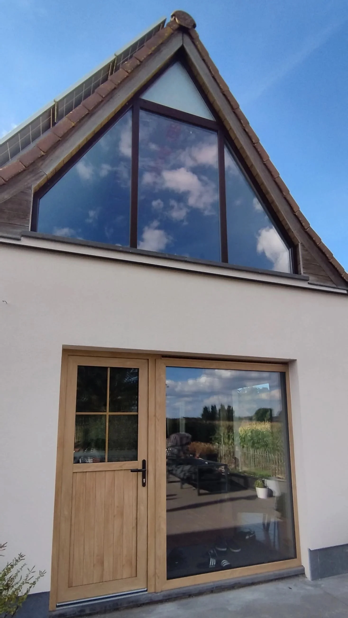 Een moderne woning met een houten voordeur en grote ramen, een zadeldak met dakpannen en zonnepanelen, en een blauwe lucht met enkele wolken.