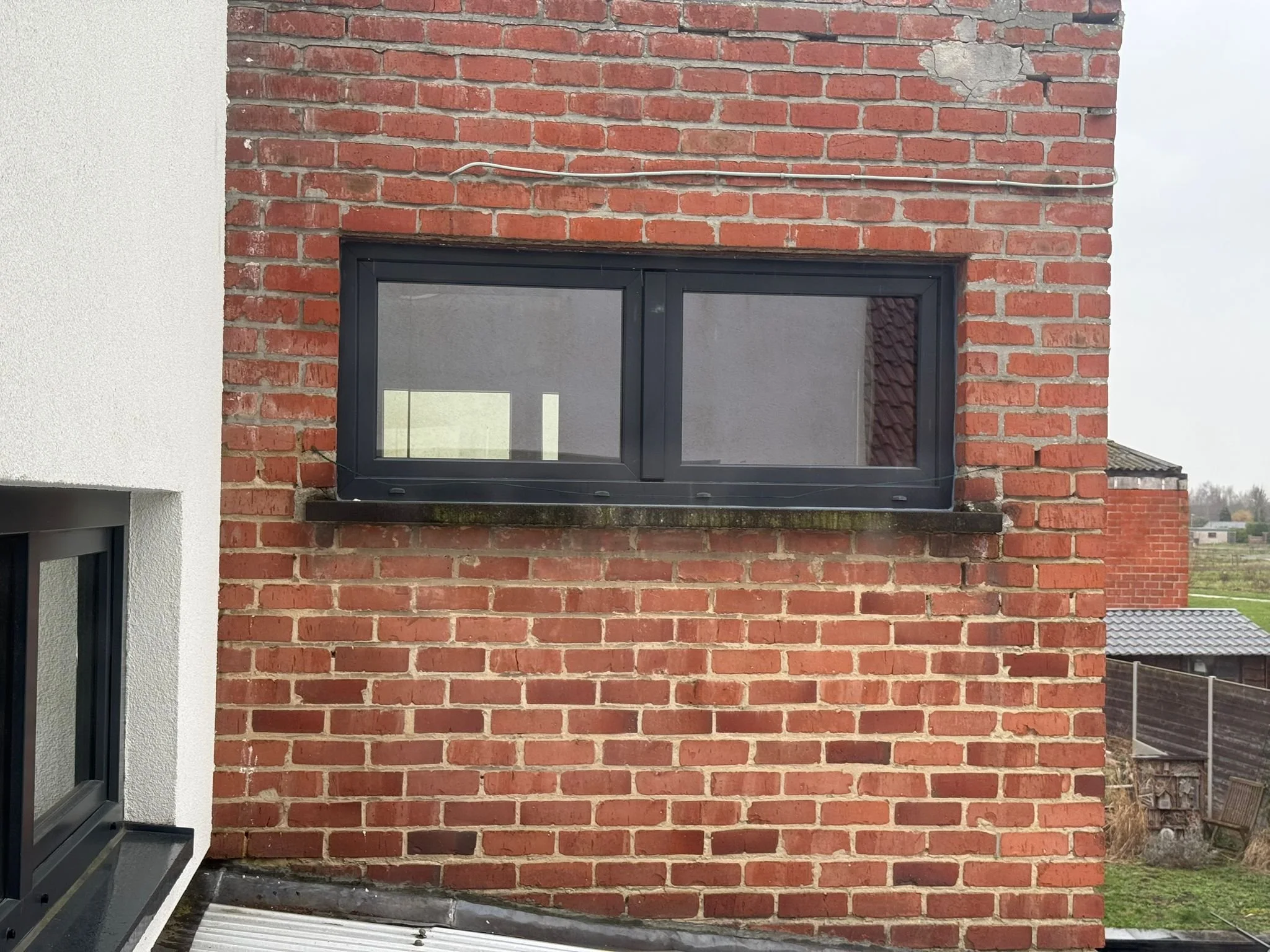 Beglazing van een gevel met roodbakstenen en een zwart kozijn, uitzicht op een tuin en andere gebouwen.