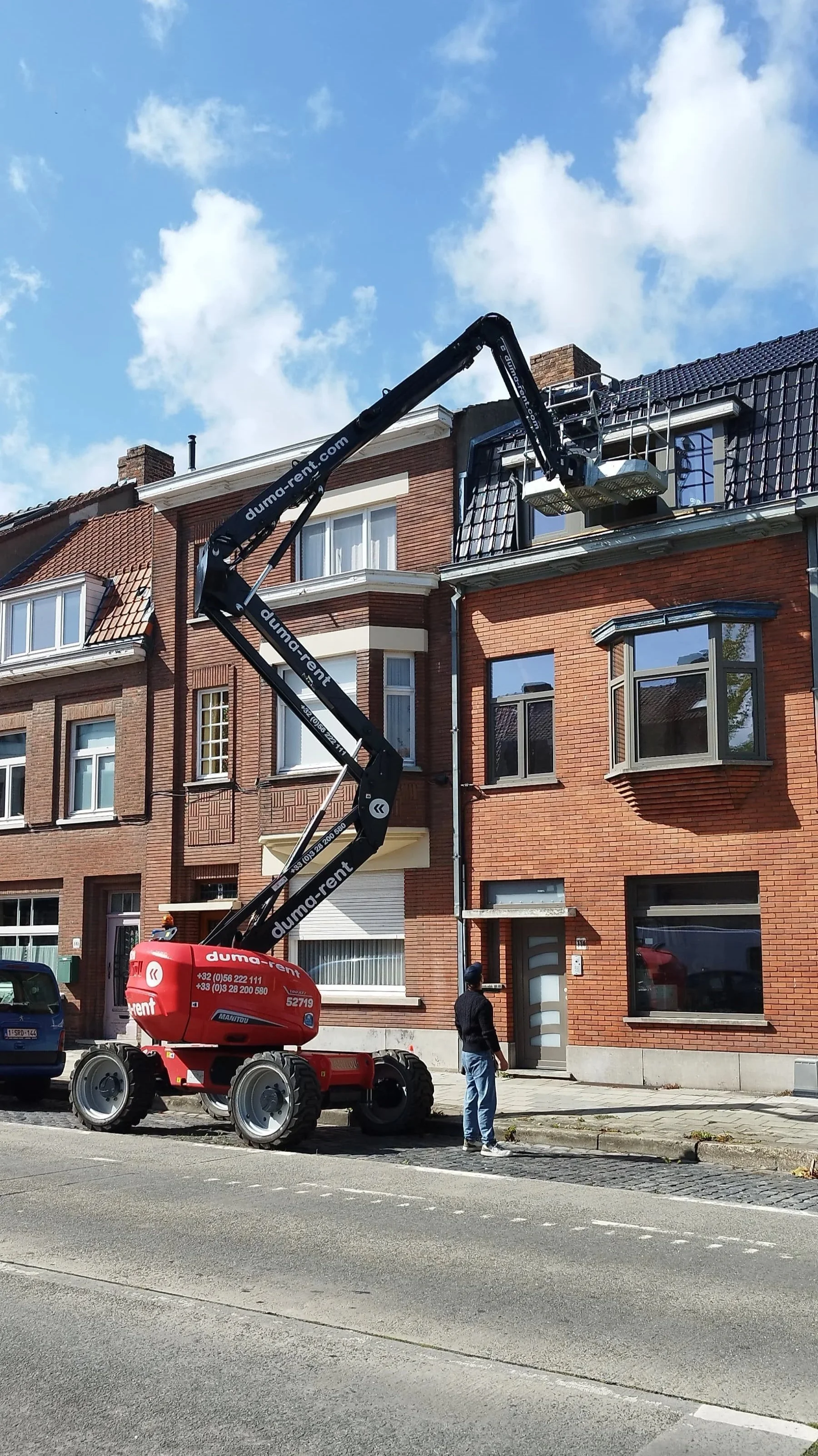 Een persoon staat op straat voor een lift die een huis op de bovenverdieping van een rood bakstenen gebouw bedient, met een blauwe lucht en wolken op de achtergrond.