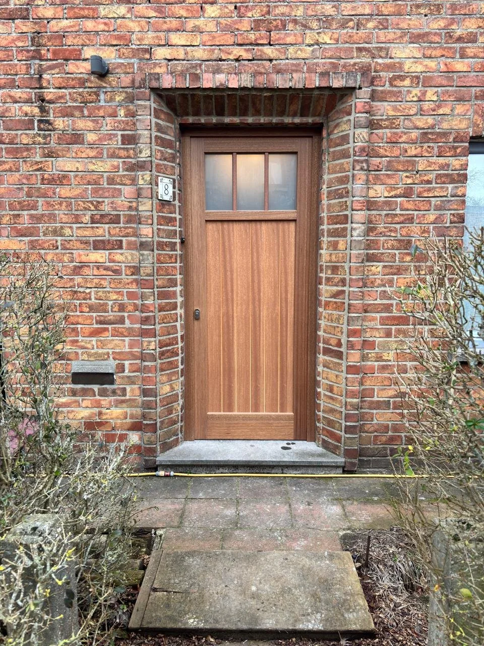 De afbeelding toont een houten voordeur op een bakstenen gevel met een huisnummer 8. Er is een kleine raam boven de deur en een kleine brievenbus links in de muur. Naast de deur staan ook twee struiken.