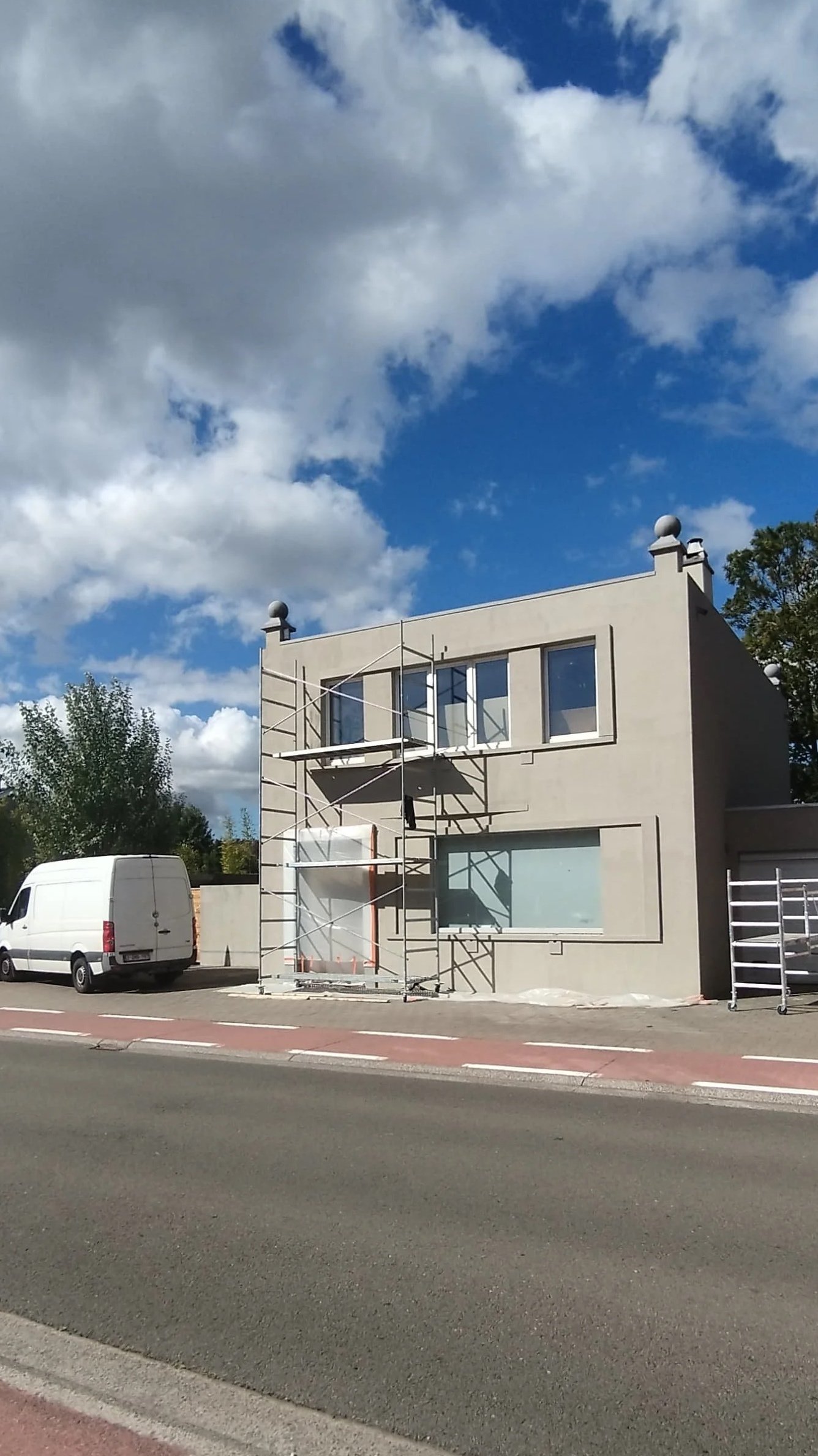 Een wit gebouw onder renovatie met verbouwing en bouwsteiger voor de gevel, naast een witte bestelbus, onder een blauwe lucht met wolken.