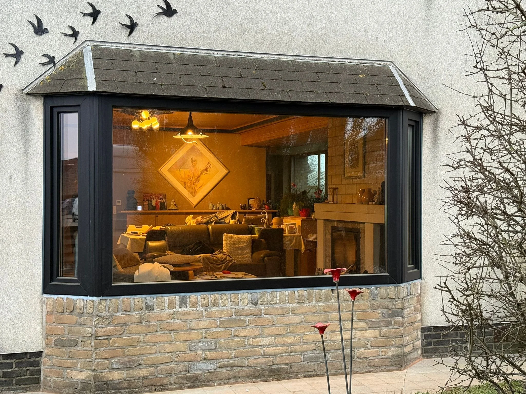 De afbeelding toont de voorkant van een huis met een grote raamuitkijk. Binnen is een gezellig interieur te zien met banken, een open haard, en wanddecoraties, verlicht door warme binnenverlichting. Buiten is een boom zonder bladeren en enkele bloemen te zien.