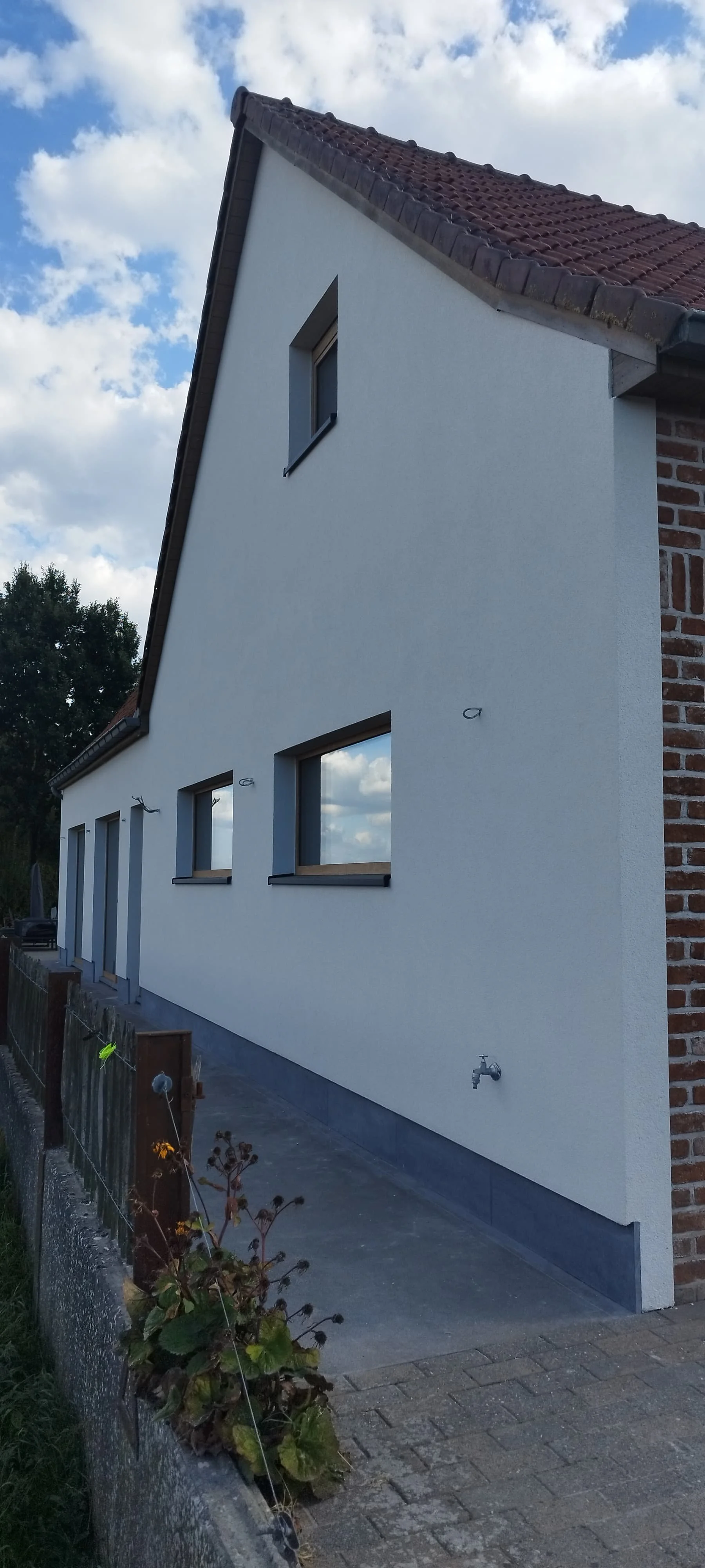 Nieuw wit huis met vier ramen, een klein dakraam en een afvoer op de zijgevel, onder een bewolkte lucht.