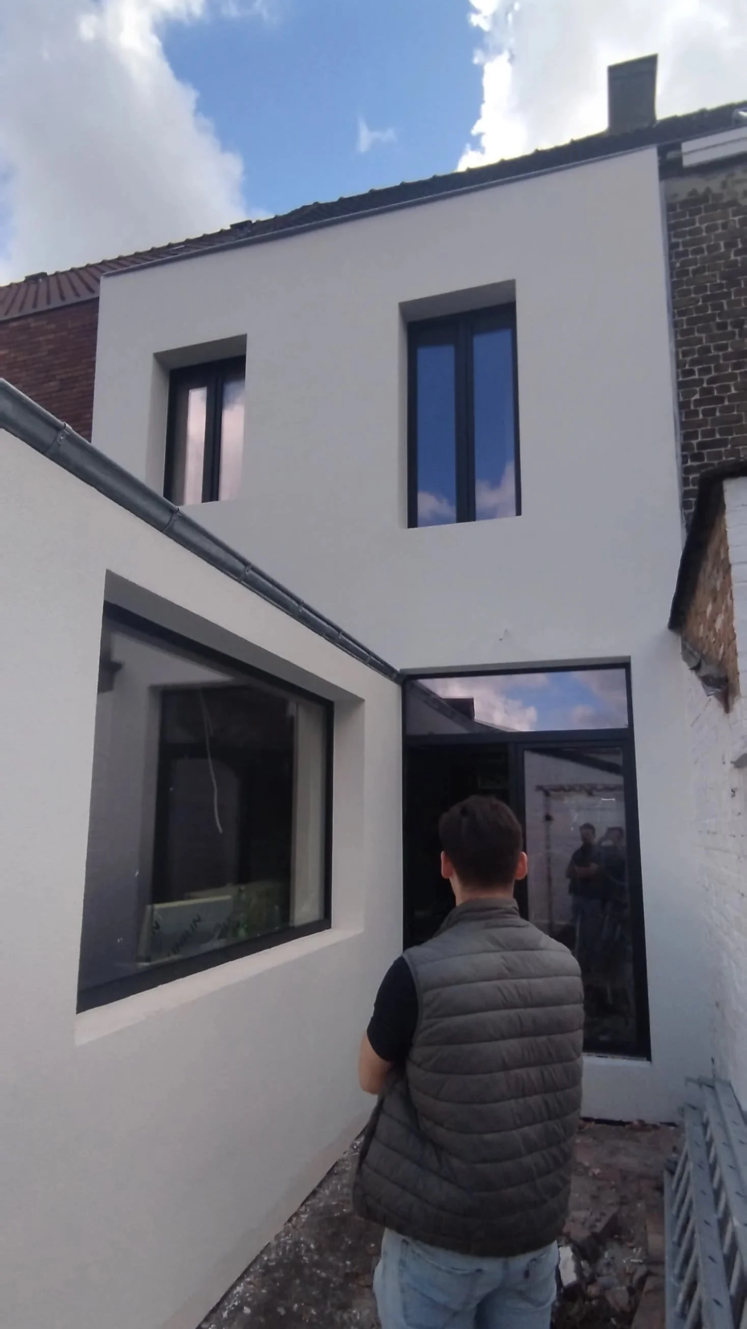 Een man staat voor een nieuwbouw wit huis met drie ramen, enkele bewolkte blauwe lucht te zien.